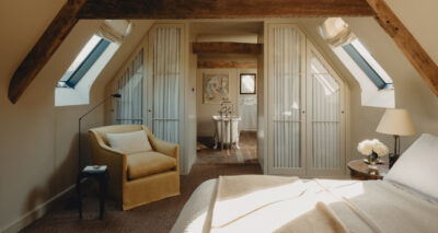 A warm, cozy attic bedroom with exposed wooden beams, skylights, a yellow armchair, a round table with flowers, and an inviting bed. Double doors open to a bright bathroom with a freestanding tub and artwork on the wall.