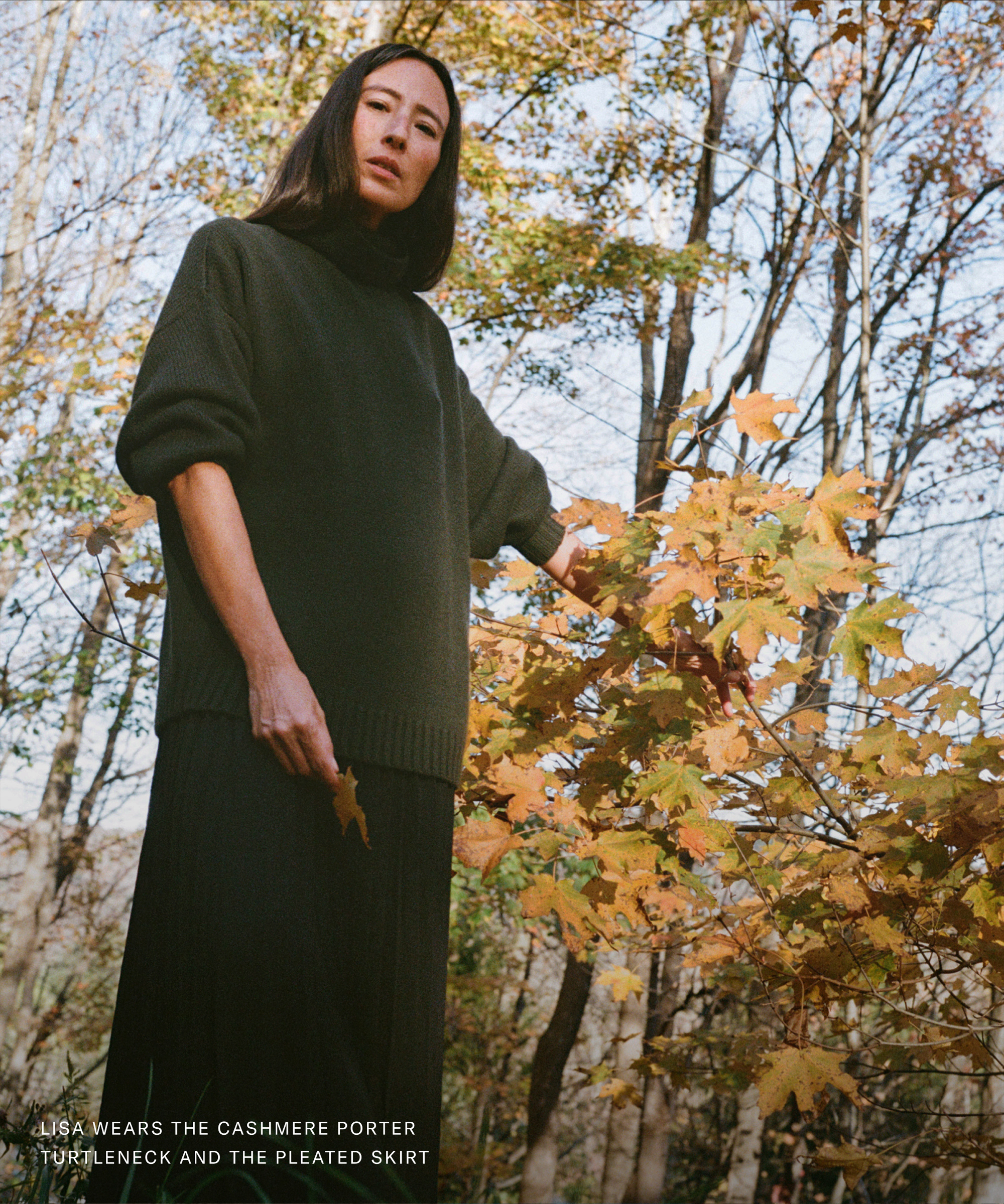 A woman stands outdoors among autumn trees, wearing a dark green turtleneck sweater and a matching pleated skirt. She looks slightly down at the camera, gently holding a branch with golden leaves, getting down to brass tacks with her effortless style.