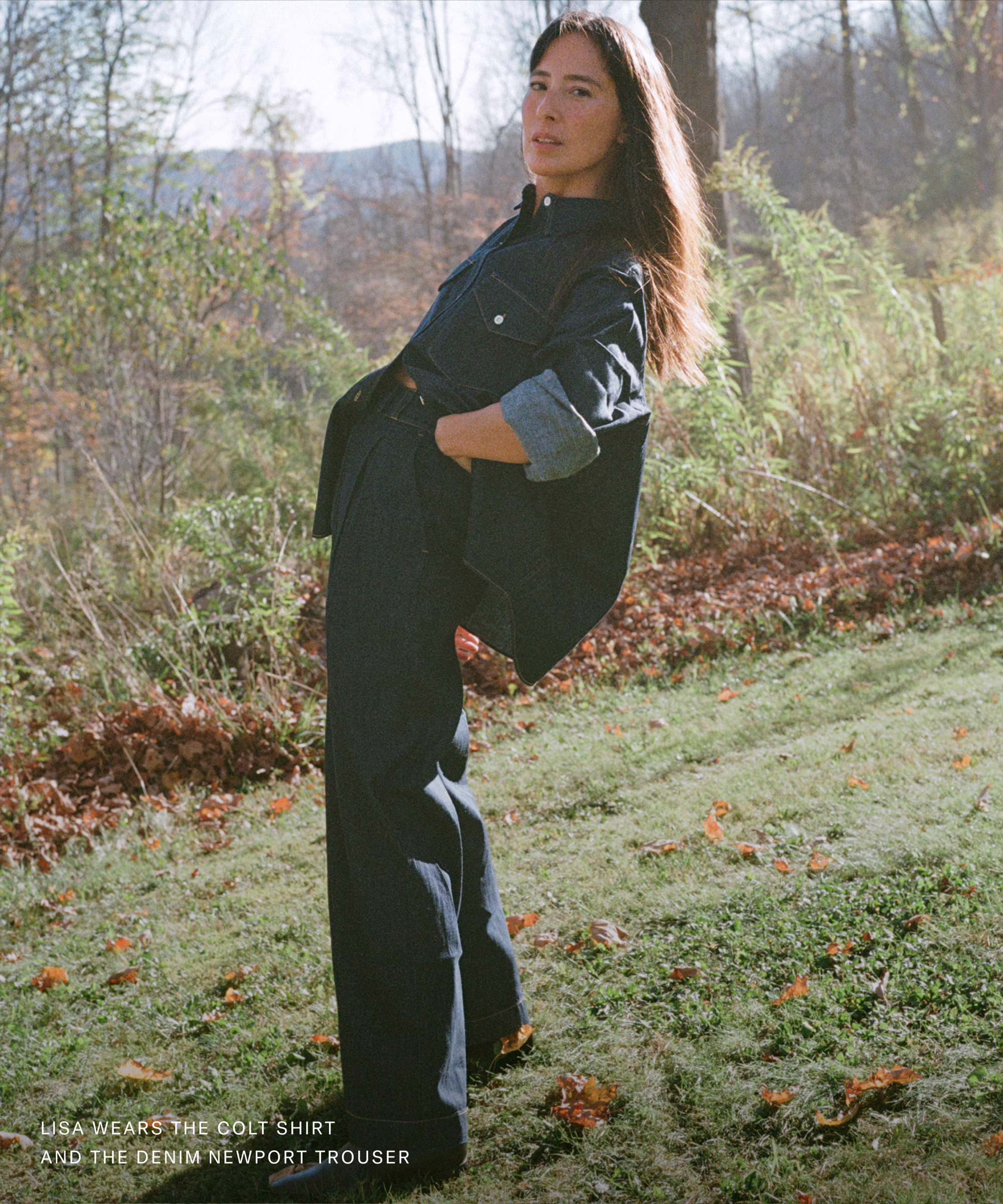 A person stands outdoors on a grassy area with trees and autumn leaves, wearing dark denim wide-leg trousers and a matching denim shirt with rolled sleeves. Sunlight highlights the natural setting, bringing things down to brass tacks in effortless style.