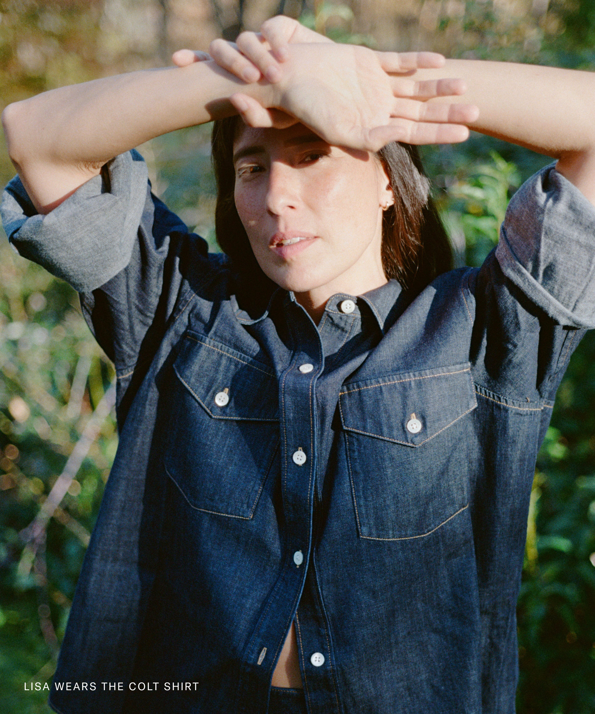 A person stands outdoors in sunlight, wearing a dark denim button-up shirt with rolled-up sleeves. Their arms are raised, partially shading their face—getting down to brass tacks. Text in the corner reads, LISA WEARS THE COLT SHIRT.