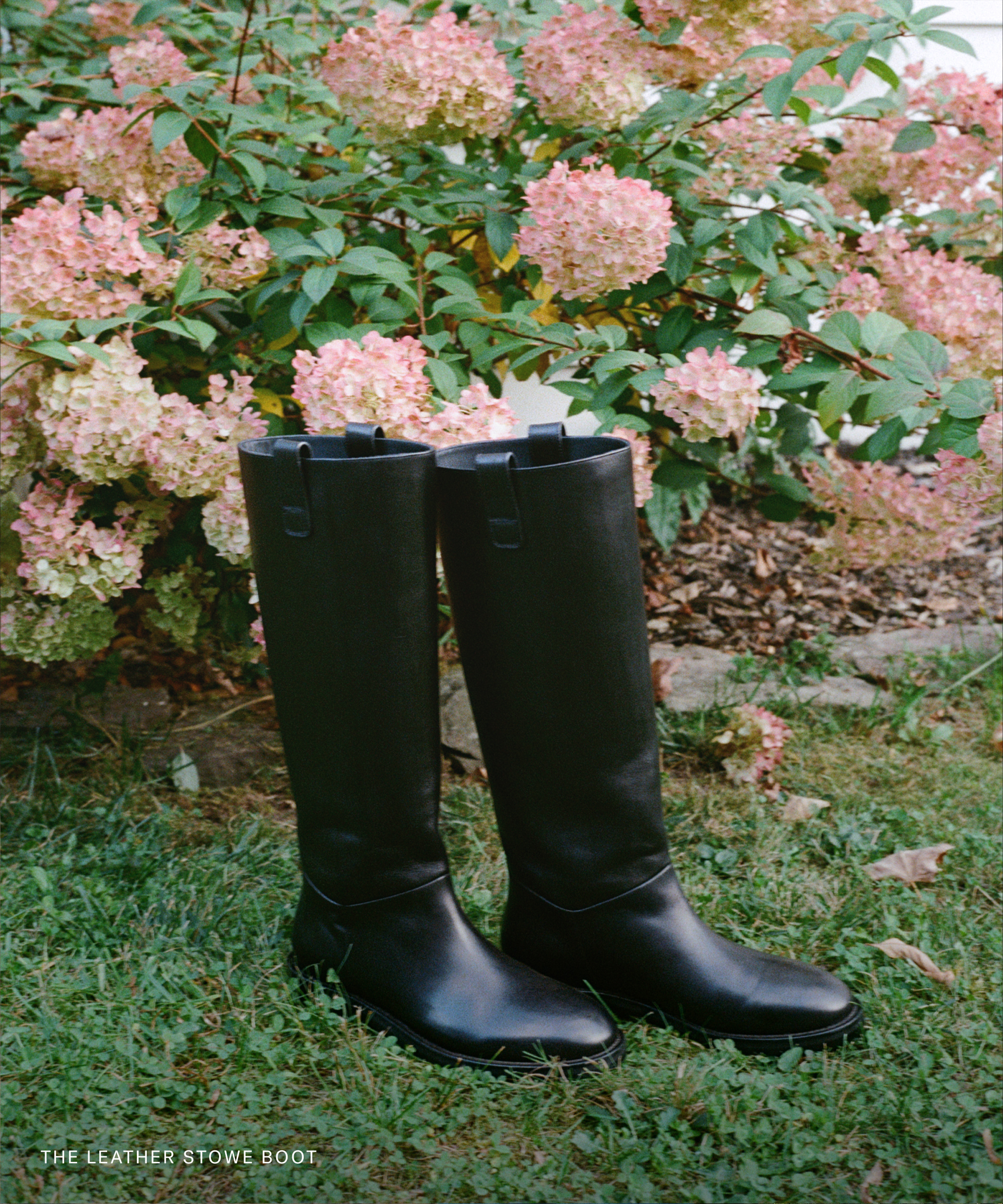 A pair of tall black leather boots stands on grass in front of blooming pink hydrangea bushes. White text at the bottom left corner reads: THE LEATHER STOWE BOOT—crafted with brass tacks for lasting durability.