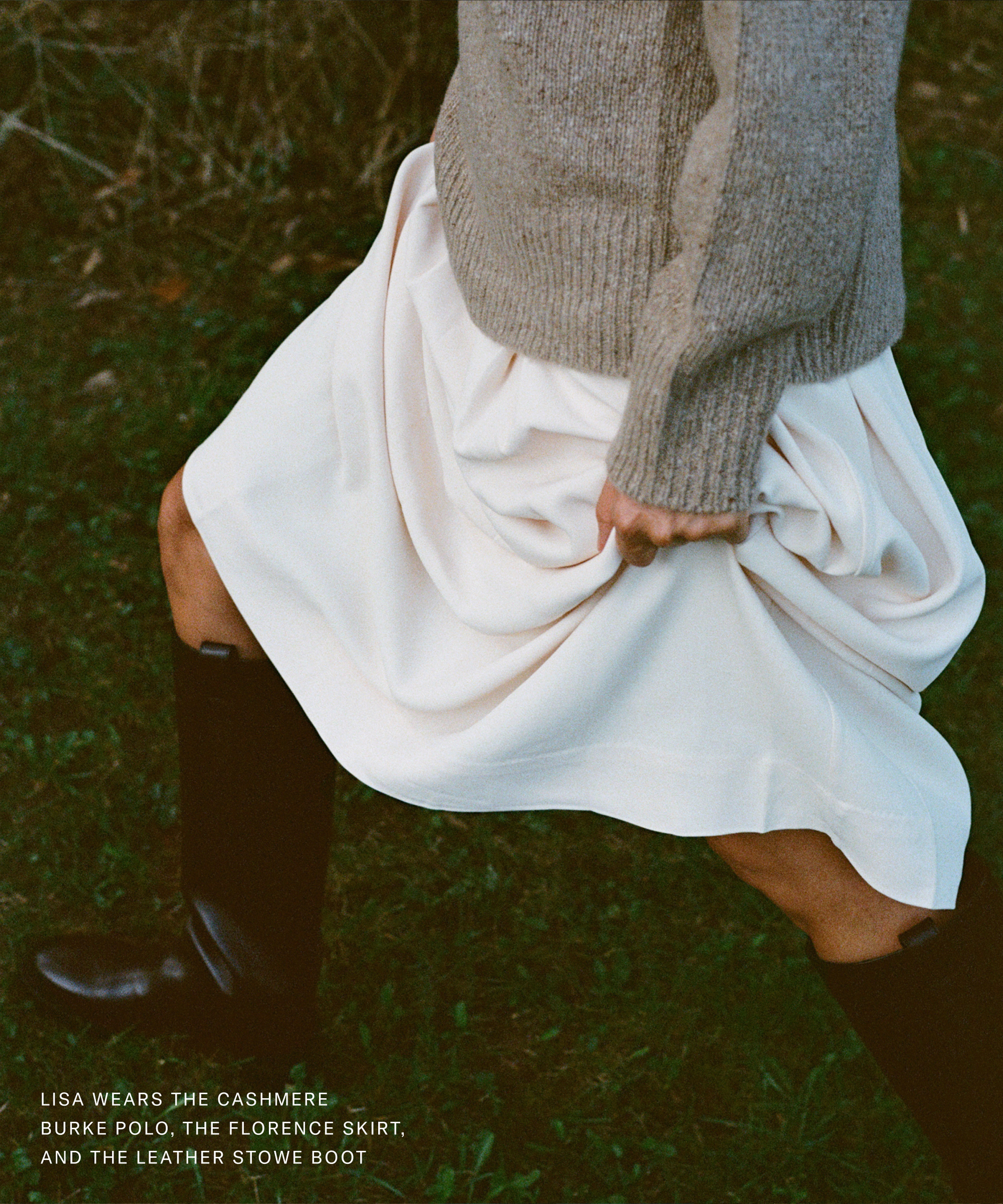 A person wearing a beige sweater, flowing white skirt, and black leather boots walks on grass. Brass tacks in the corner detail the outfit: cashmere polo, Florence skirt, and leather Stowe boot.