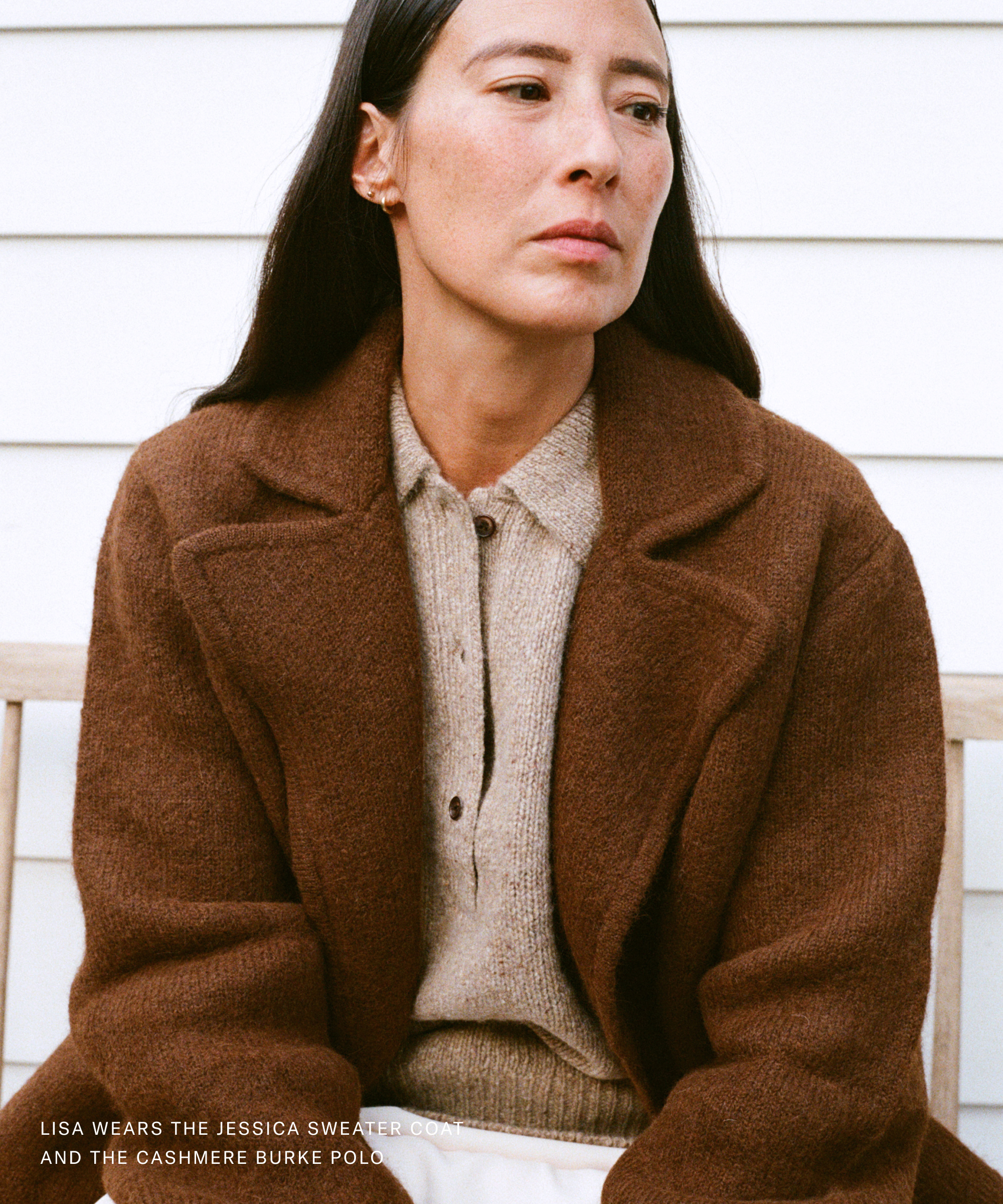A woman with long dark hair sits on a bench, wearing a brown coat over a beige sweater, looking thoughtfully to the side. The background is white siding. Text in the corner gets down to brass tacks, detailing her outfit.