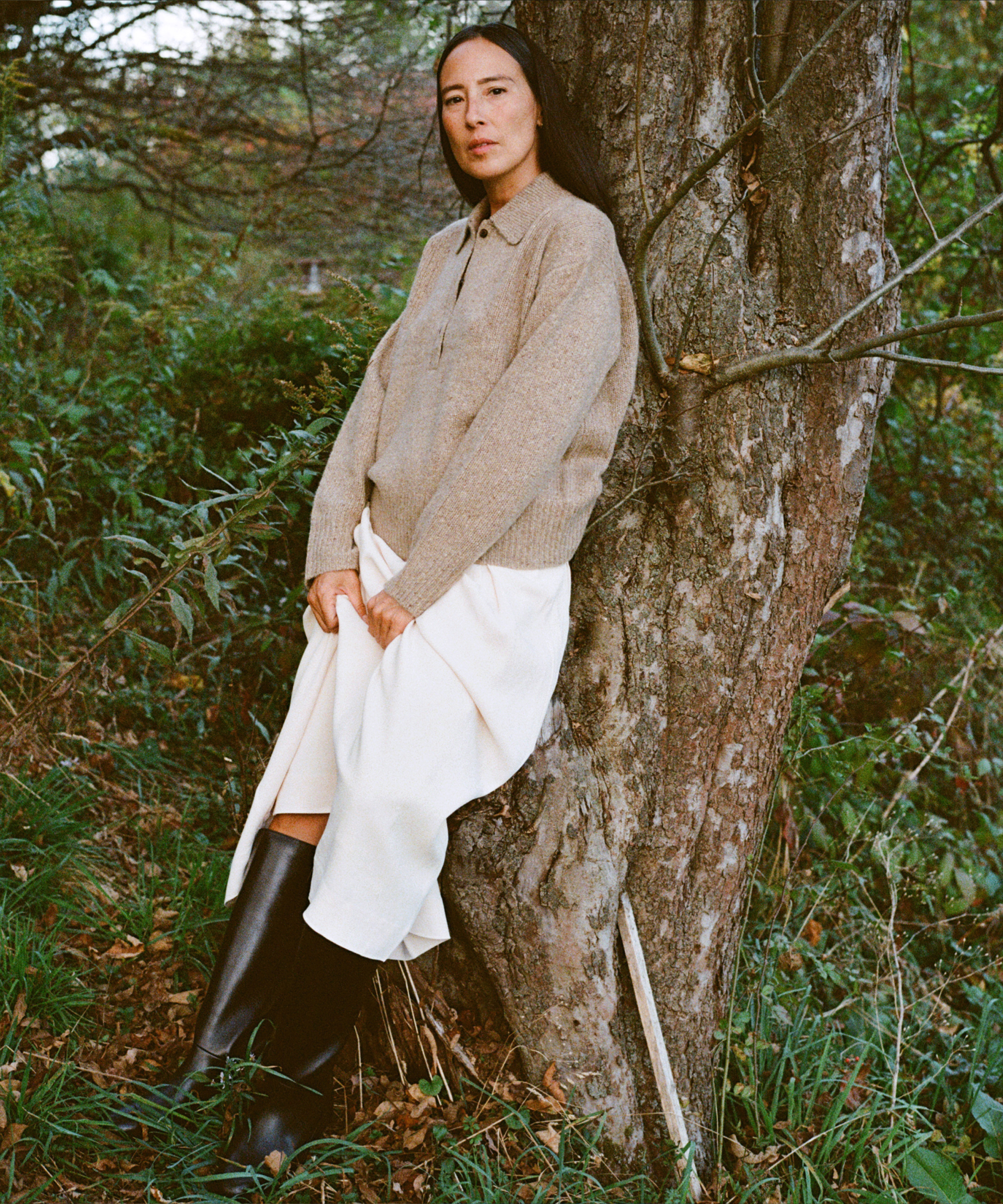 A person with long dark hair leans against a tree in an outdoor, wooded area. Getting down to brass tacks, they wear a tan sweater, light beige skirt, and tall black boots among green foliage and fallen leaves.