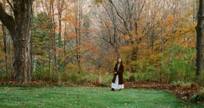 A woman in a long brown coat and white dress stands on green grass, surrounded by tall trees with colorful autumn leaves—getting down to brass tacks in this peaceful, wooded area.
