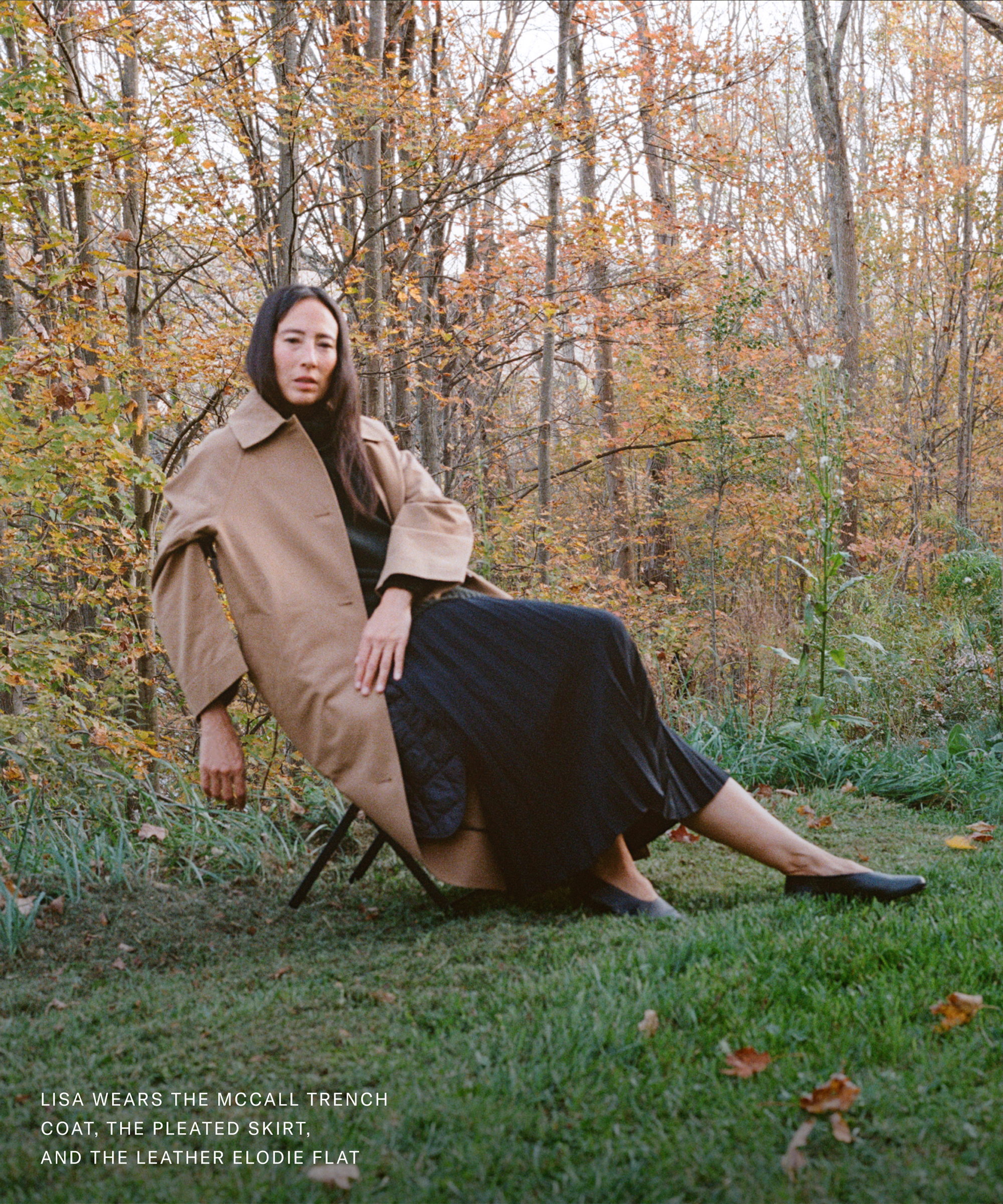 A woman sits on a chair outdoors in autumn, getting down to brass tacks in a tan trench coat over a long black pleated skirt and black flats. Trees with fall leaves and green grass set the scene in the background.