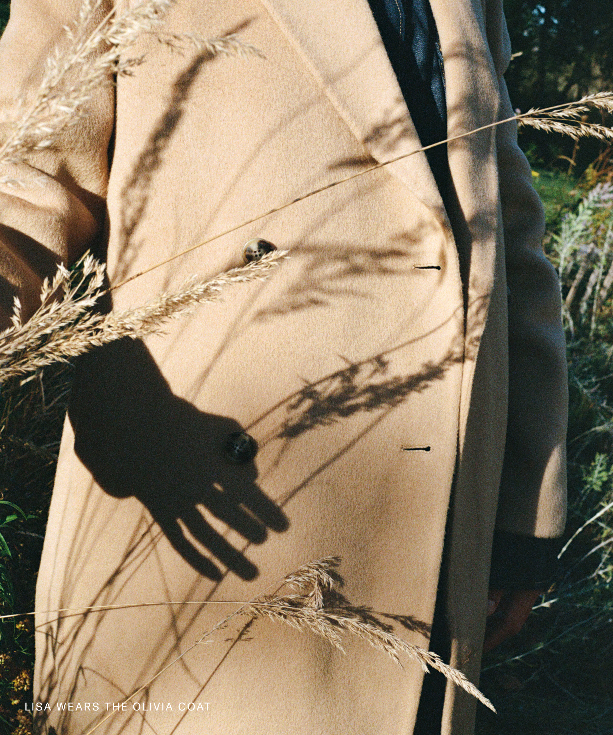 A person wearing a beige coat stands among tall grasses. Sunlight casts a distinct shadow of their hand on the coat, getting down to brass tacks. Text in the corner reads, “LISA WEARS THE OLIVIA COAT.”.