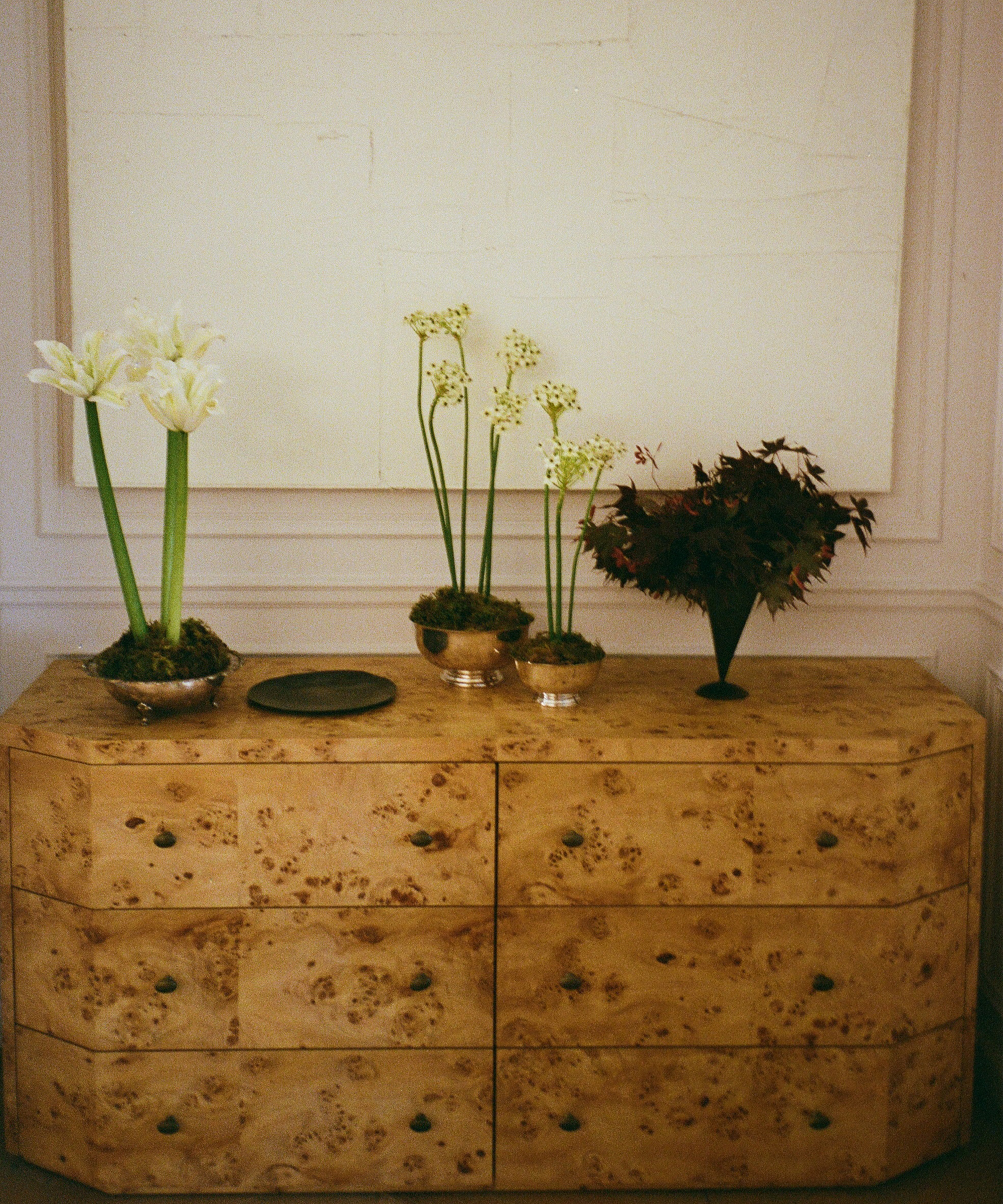 A light wood dresser with green knobs holds vases of white flowers and dark foliage, a small bowl, and a plate. A large blank white frame hangs on the wall behind it.