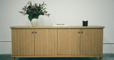 A light wooden cabinet with four doors stands against a white wall. On top, there is a vase of flowers on the left, a closed book in the center, and a lit black candle on the right.