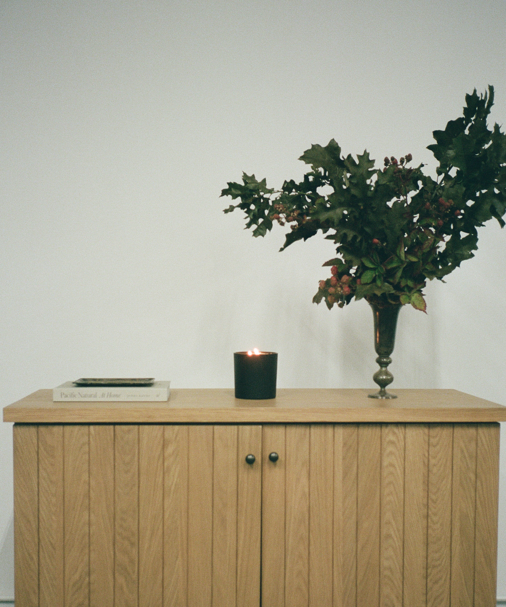 A wooden cabinet with vertical grooves holds a closed book, a lit black candle, and a vase with leafy branches and berries, set against a plain white wall.