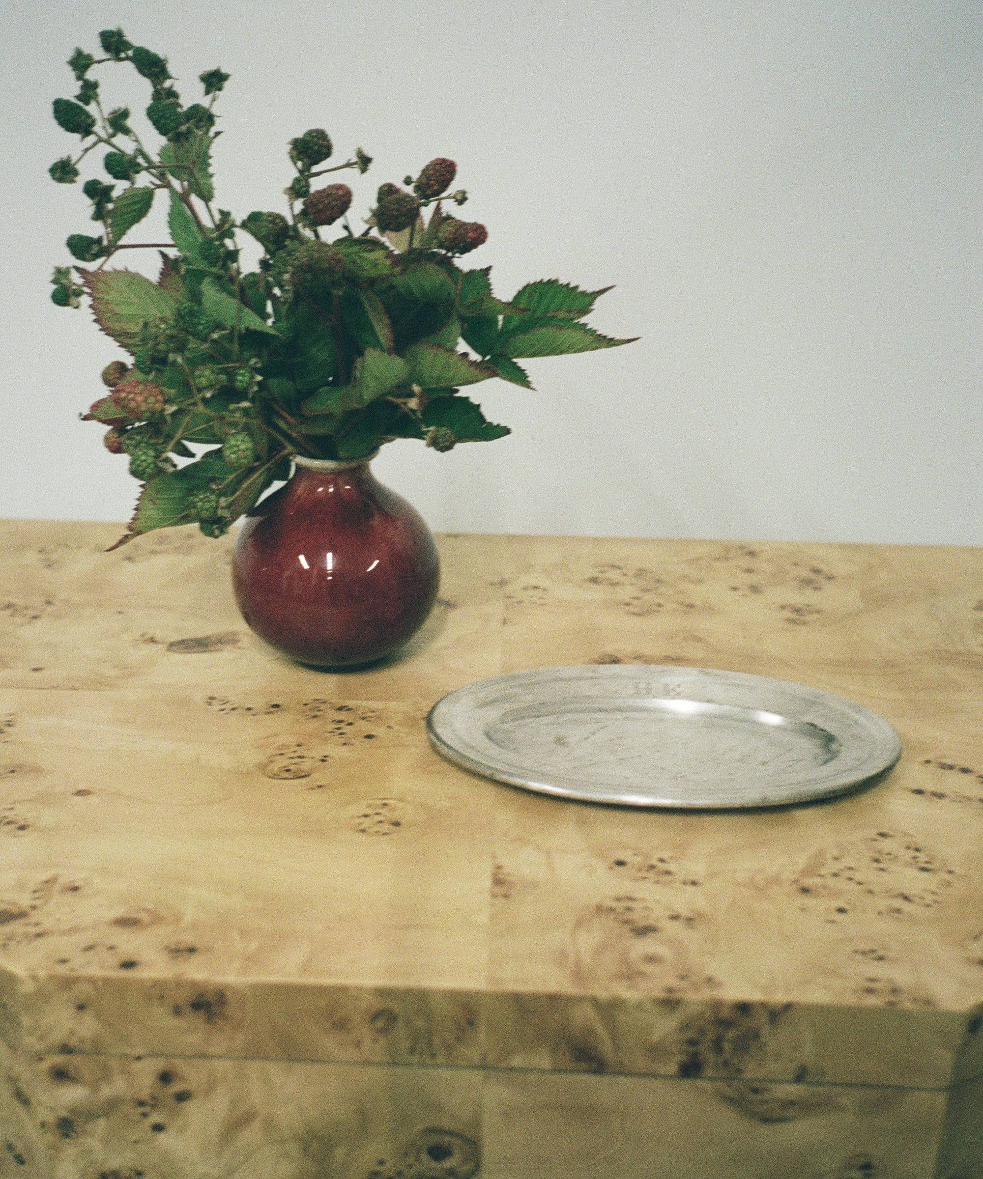 A red vase with green leafy branches and berries sits on a light wood table next to a round, silver metal plate against a plain white background.