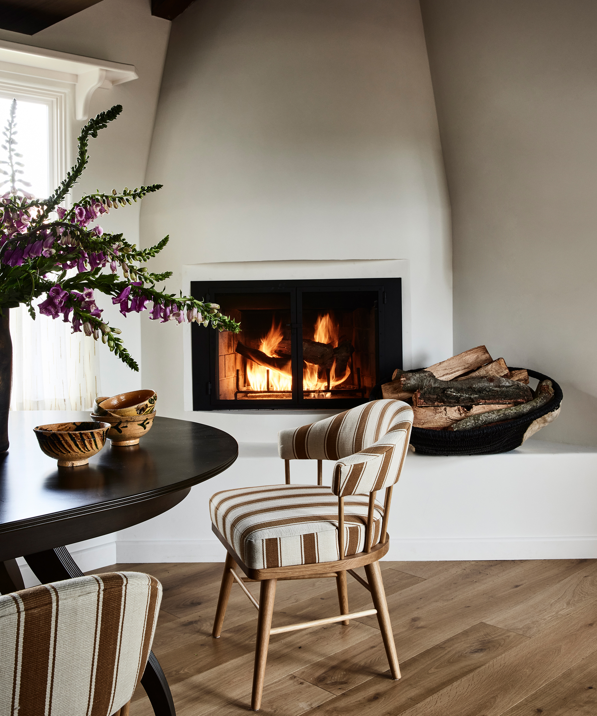 A cozy Santa Ynez room with a modern fireplace burning wood, a basket of firewood beside it, a round dark table with striped chairs, and a vase of purple flowers—all set on light wooden floors.