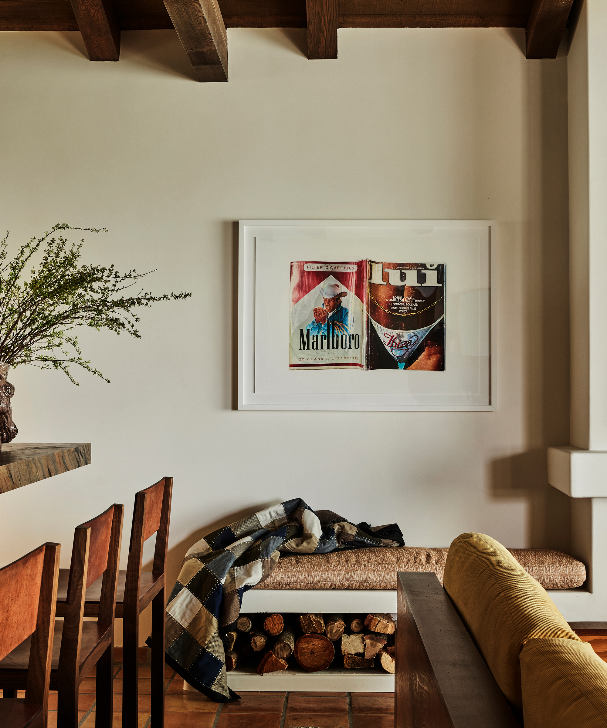 A cozy Santa Ynez living space with a wooden bench, plaid blanket, firewood storage underneath, and framed artwork above featuring vintage Marlboro and Budweiser ads. Wooden chairs and a vase with greenery complete the scene.