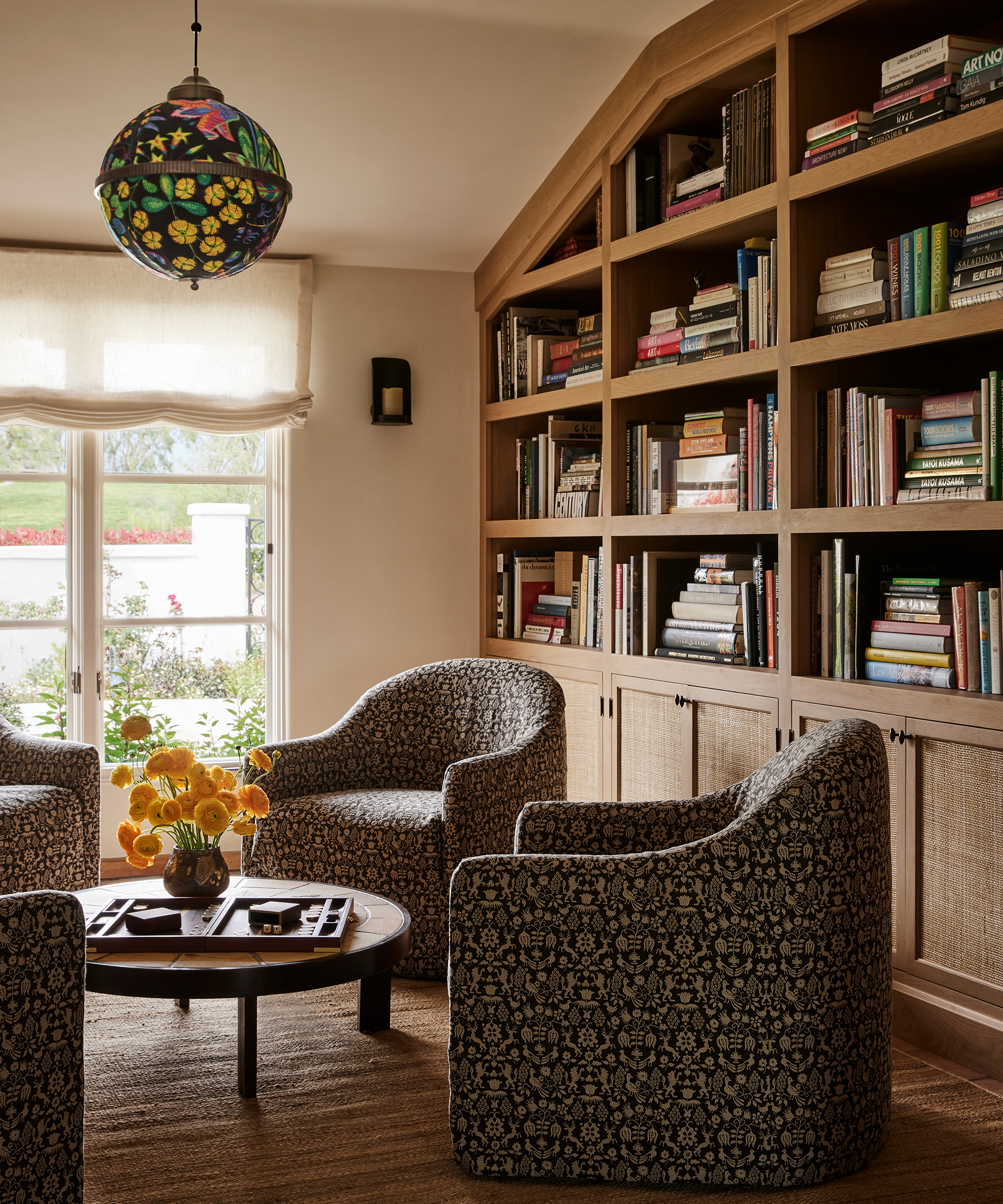 A cozy Santa Ynez reading nook features patterned armchairs around a circular coffee table with yellow flowers, set beside large windows and a built-in bookshelf filled with books. A colorful globe-shaped pendant light hangs from the ceiling.