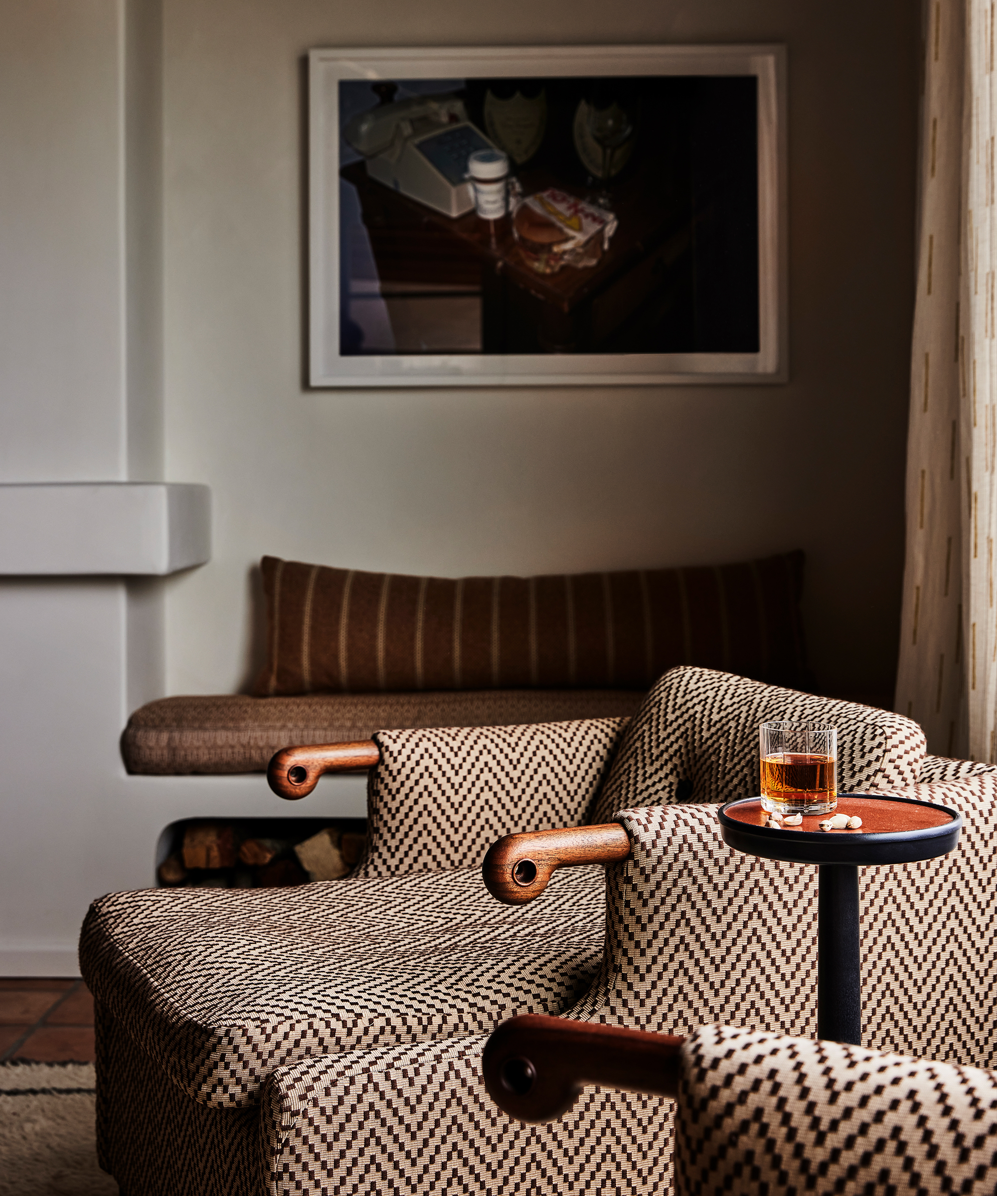 Two patterned armchairs and a small round table with a glass of whiskey and nuts create a cozy, softly lit Santa Ynez retreat. A striped bench and framed artwork complete the inviting scene in the background.