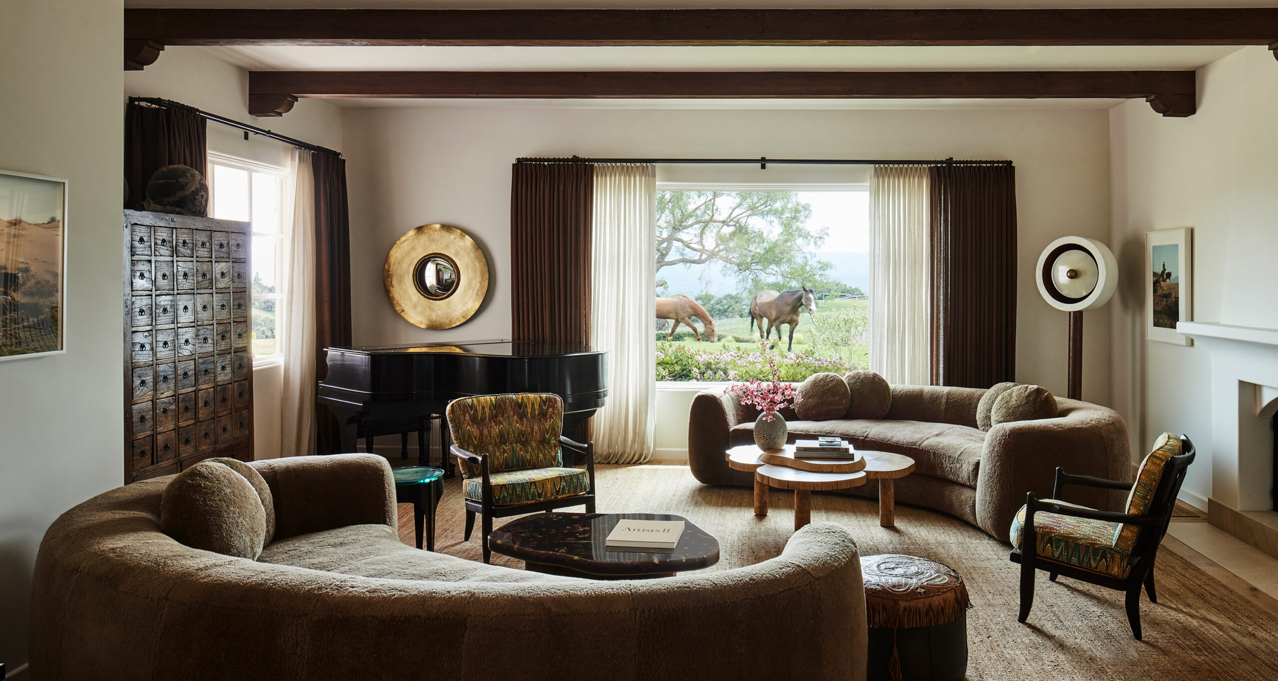 A stylish Santa Ynez living room with curved sofas, a grand piano, and a large window showcasing a scenic outdoor view with two horses grazing near a tree. Warm, neutral tones and wooden beams create a cozy atmosphere.