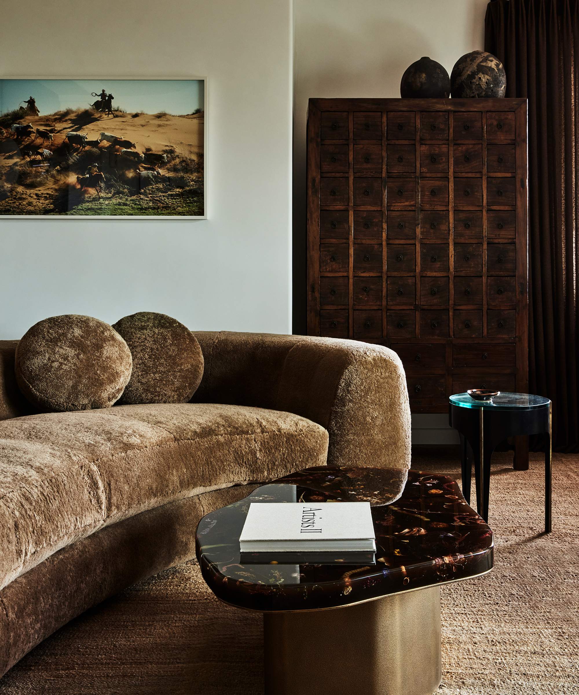 Cozy living room with a plush brown curved sofa, round pillows, a dark marble coffee table with books, a vintage wooden cabinet, and wall art of a Santa Ynez desert scene. Warm, earthy tones dominate the decor.
