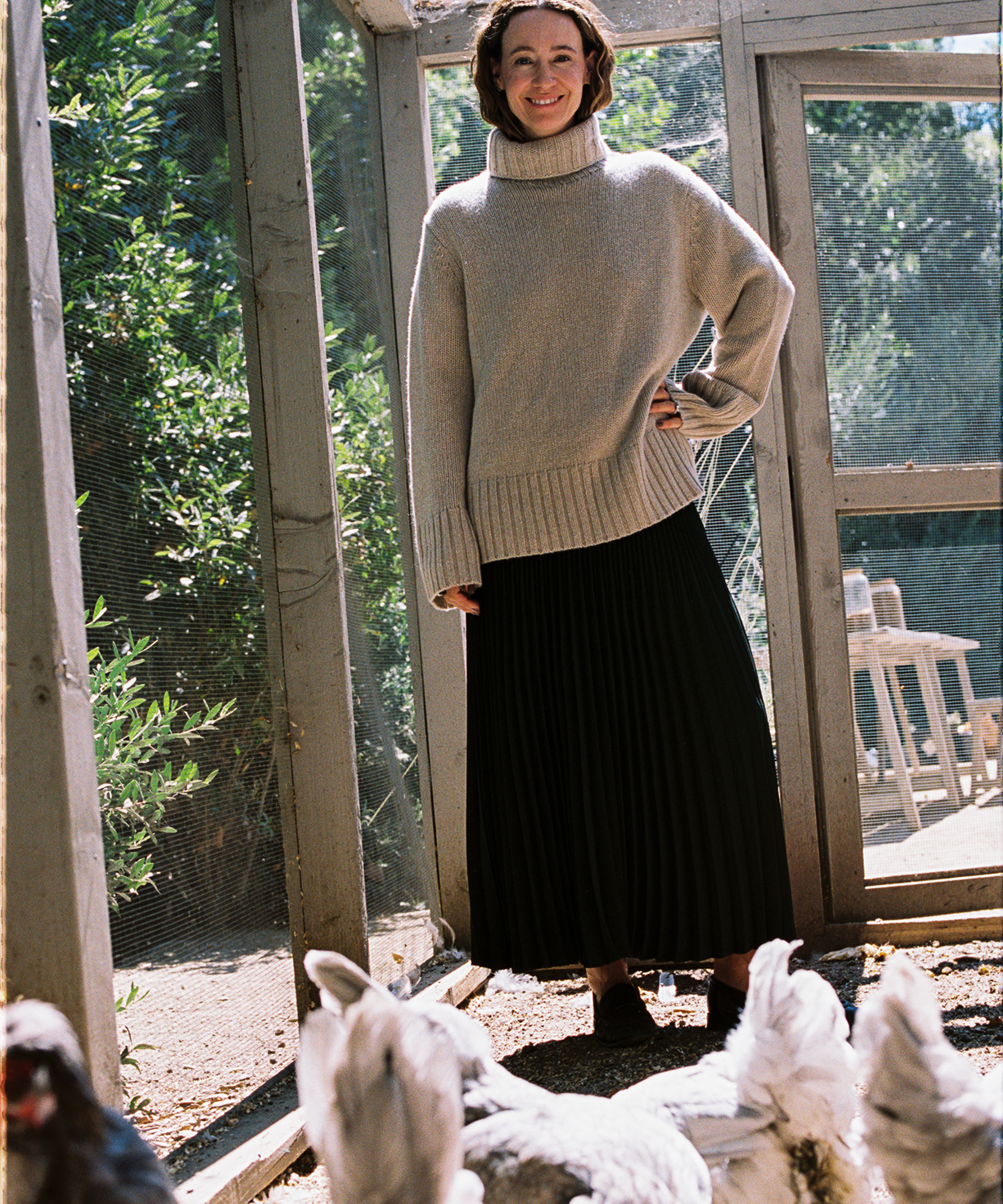A woman wearing a light turtleneck sweater and long dark skirt stands in a sunlit chicken coop, smiling with one hand on her hip. Chickens are gathered near her feet, and greenery is visible outside the enclosure.