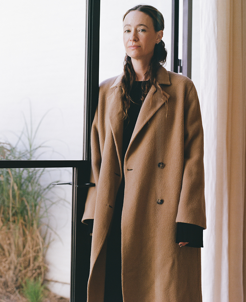 A woman with long brown hair stands indoors near a glass door, wearing a long brown overcoat over dark clothing. Light filters in, highlighting her face; plants and curtains are visible in the background.