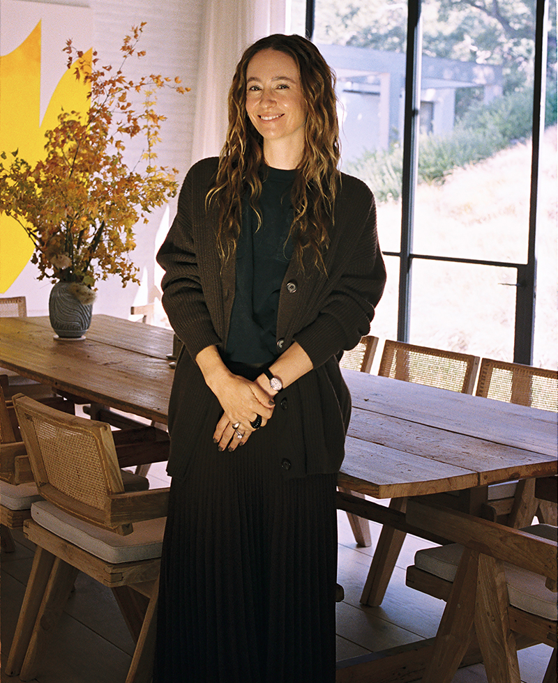 A woman with long wavy hair stands smiling by a wooden dining table with wicker chairs, wearing a dark cardigan and skirt. A vase with yellow flowers is on the table, and large windows let in natural light.