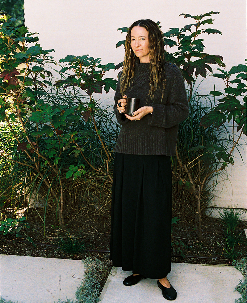 A woman with long wavy hair stands outside in front of leafy plants, wearing a loose dark sweater, a long black skirt, and black shoes, holding a mug and looking at the camera with a soft smile.