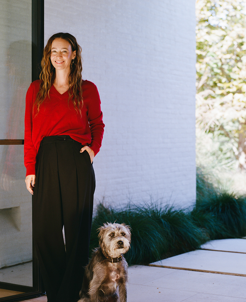 A woman wearing a red sweater and black pants stands smiling outside next to a gray, scruffy dog. They are in front of a glass door with white walls and greenery in the background.