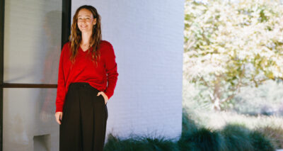 A woman with long brown hair wearing a red sweater and black pants stands outside near a white building, smiling with one hand in her pocket. Sunlight and greenery are visible in the background.