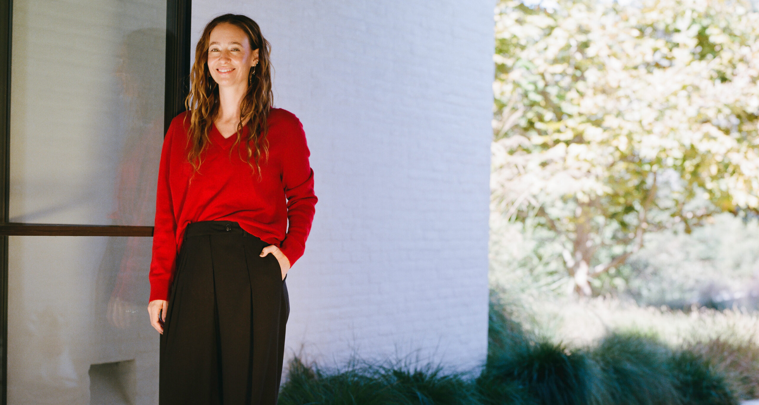 A woman with long brown hair wearing a red sweater and black pants stands outside near a white building, smiling with one hand in her pocket. Sunlight and greenery are visible in the background.
