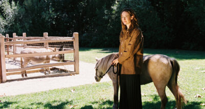 A woman with long brown hair stands on grass holding the leash of a small pony. Behind her, two donkeys are in a wooden enclosure. Trees and sunlight fill the background.