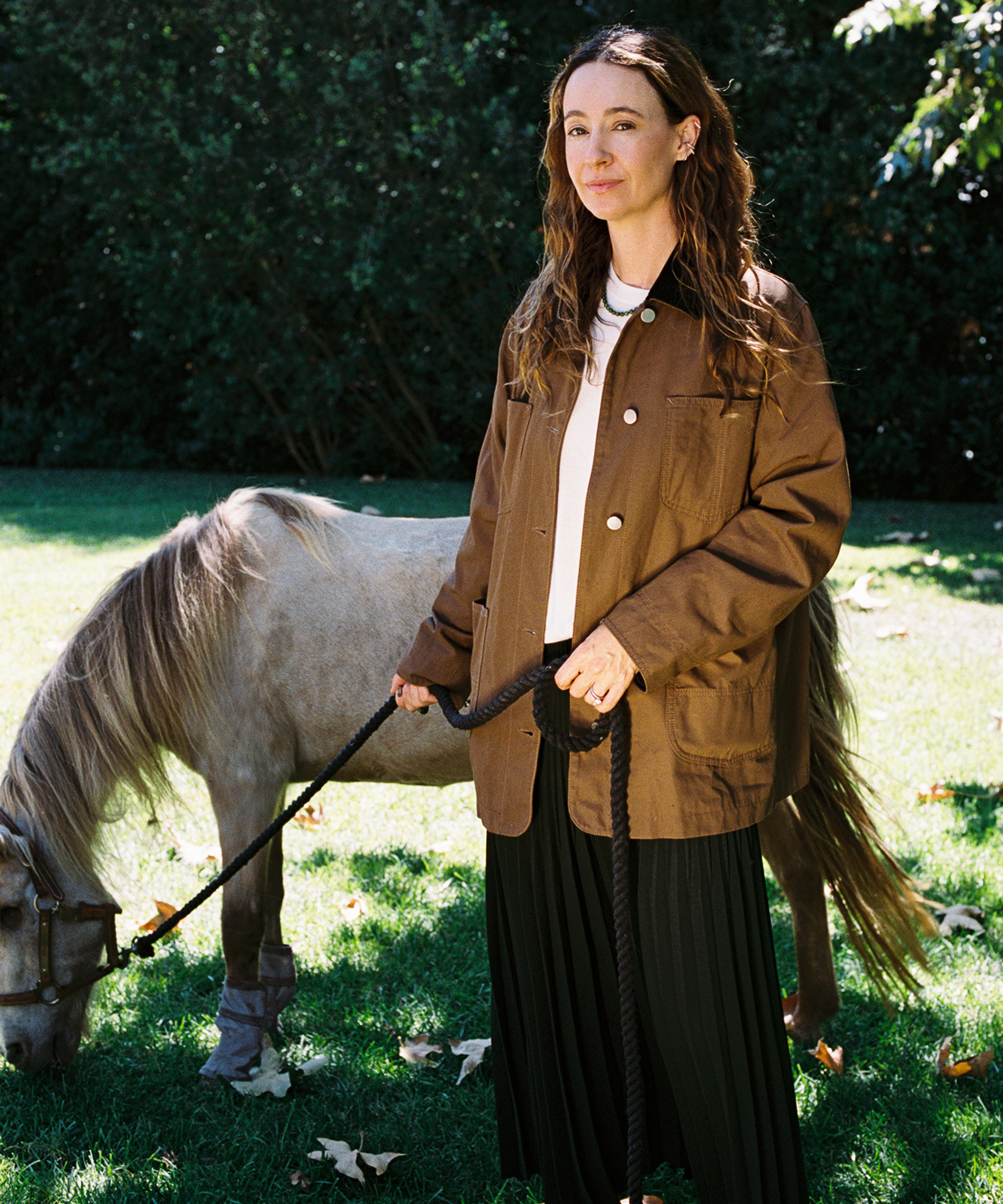 A woman with wavy hair wearing a brown jacket stands on grass, holding the lead of a small horse that is grazing. Trees and greenery are visible in the background.