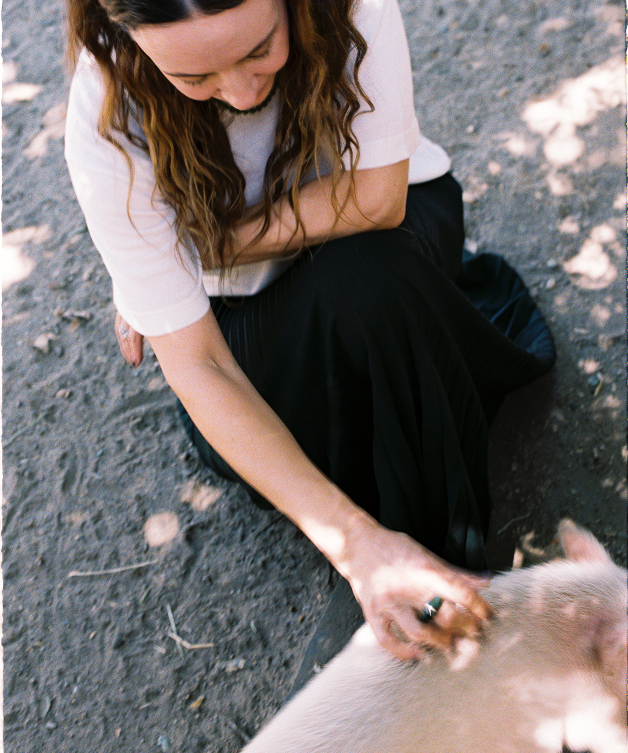 A person with wavy brown hair, wearing a white top and black skirt, sits on the ground and gently pets a light-colored animal outdoors, with dappled sunlight on the dirt below.