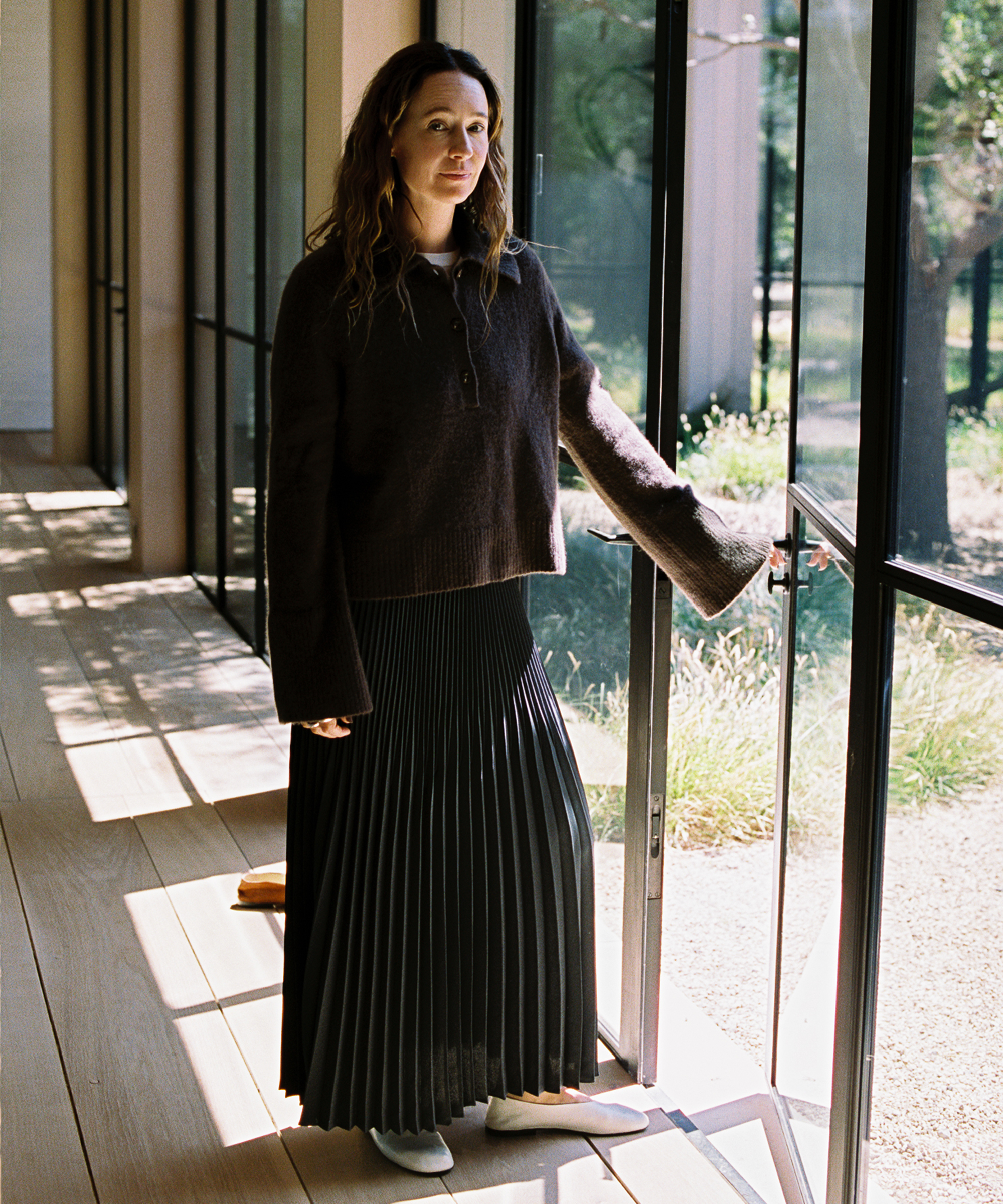 A woman with long hair wearing a dark sweater and a pleated skirt stands indoors by a glass door, with sunlight streaming in and greenery visible outside.