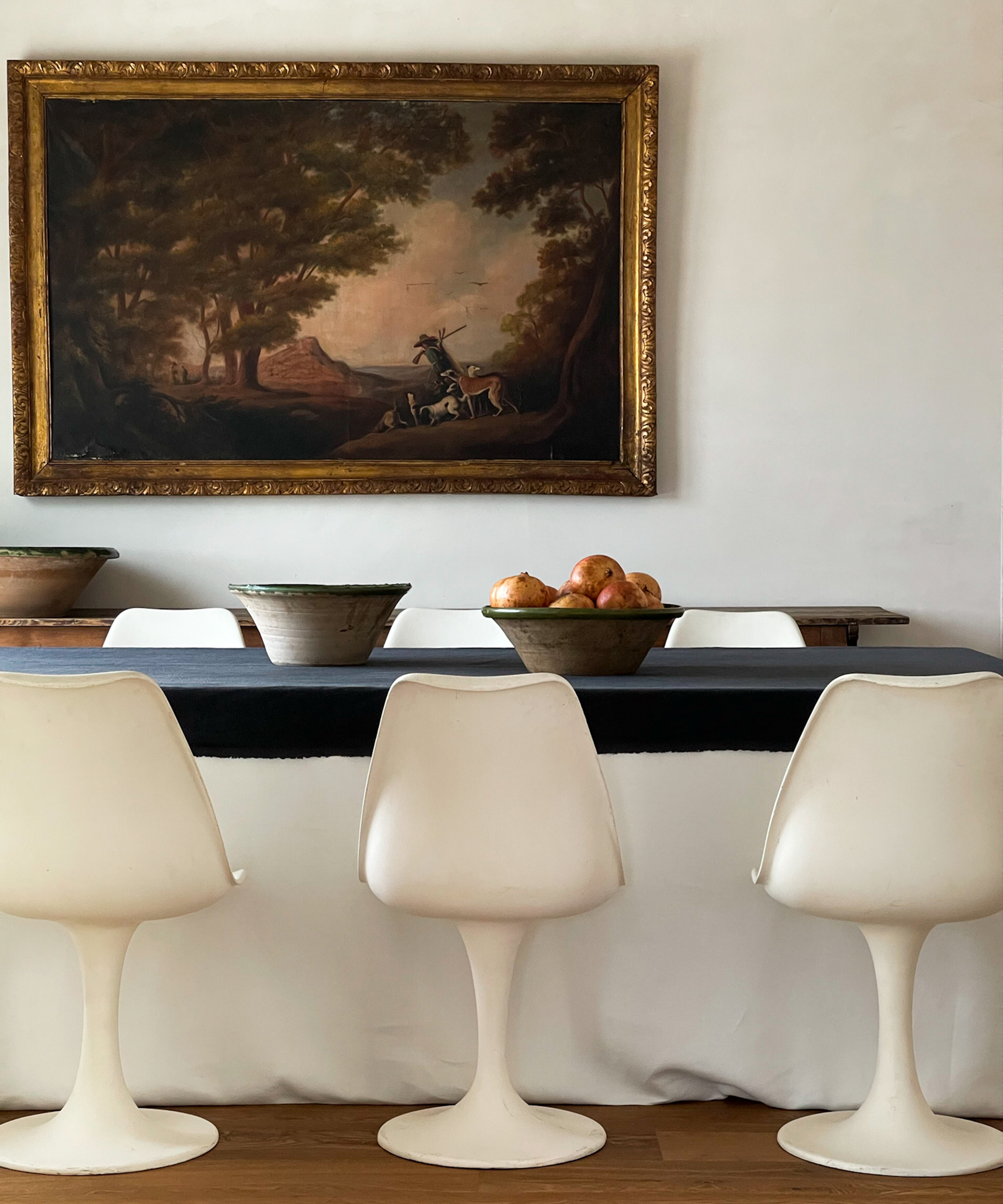 A dining table with four white modern chairs, a dark tablecloth, a bowl of pomegranates, two bowls, and a large framed painting of a horseback scene on the wall behind.