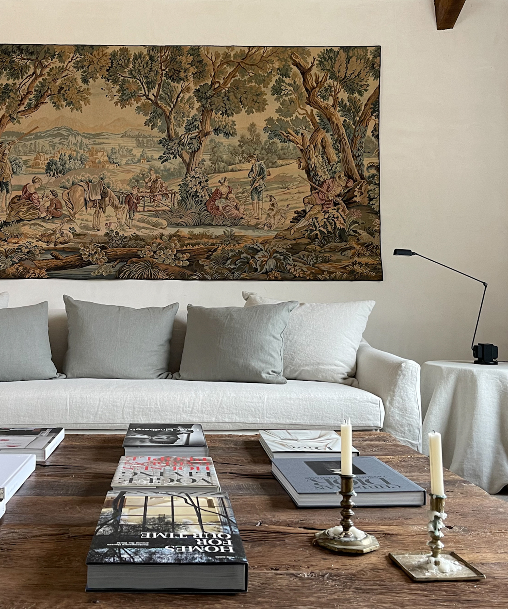 A cozy living room with a white sofa, light grey pillows, and a large tapestry depicting a nature scene on the wall. A wooden coffee table holds books and two candlesticks; a black lamp sits on a side table.