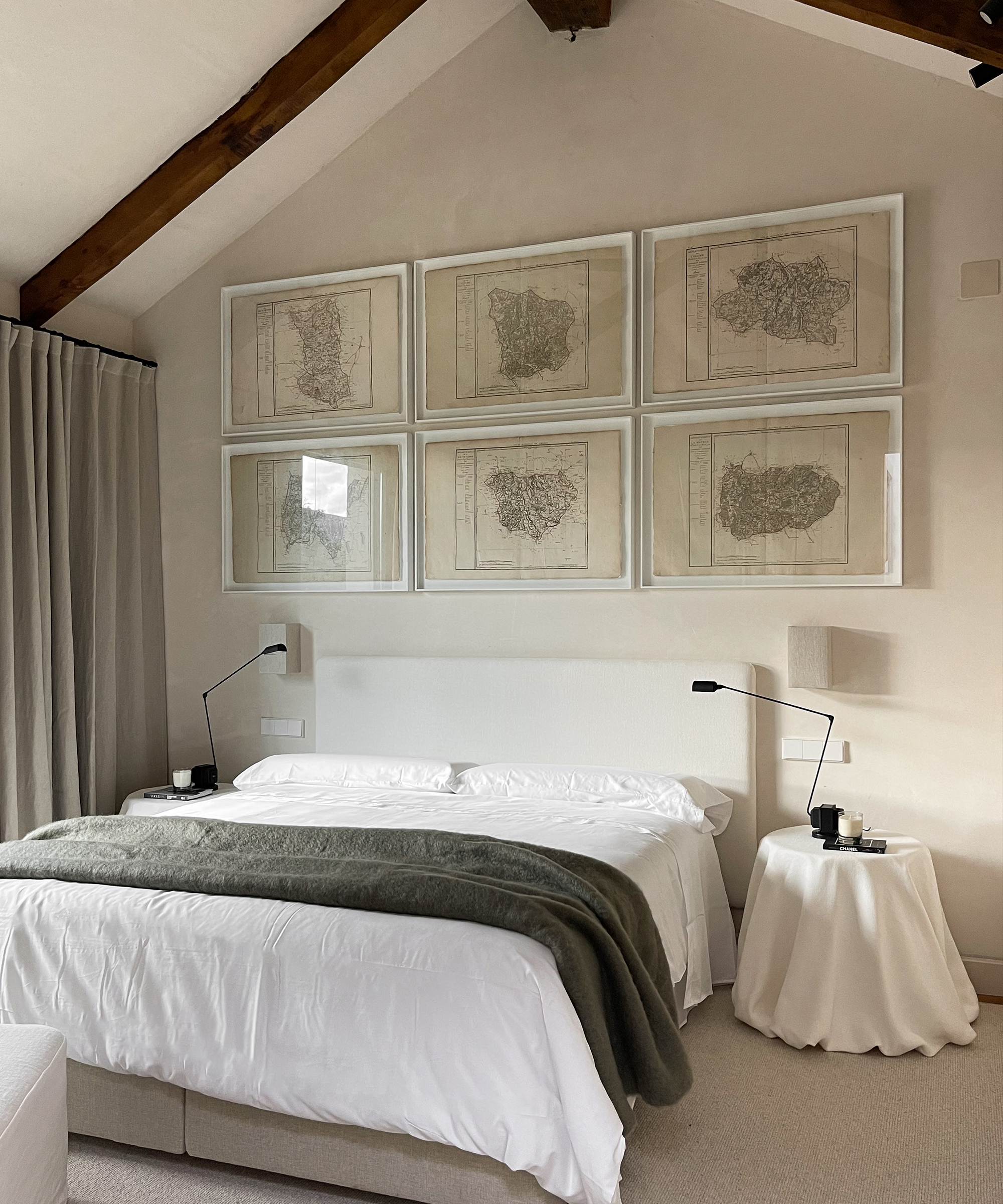 A modern bedroom with a white bed, gray blanket, round bedside tables, and black lamps. Six framed map prints hang on the light-colored wall above the bed. The room has sloped ceilings with exposed wooden beams.