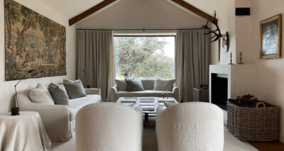 A cozy living room with neutral tones, featuring two white armchairs, a white sofa, a wooden coffee table, a large window with beige curtains, a fireplace with a basket of firewood, and a wall tapestry.