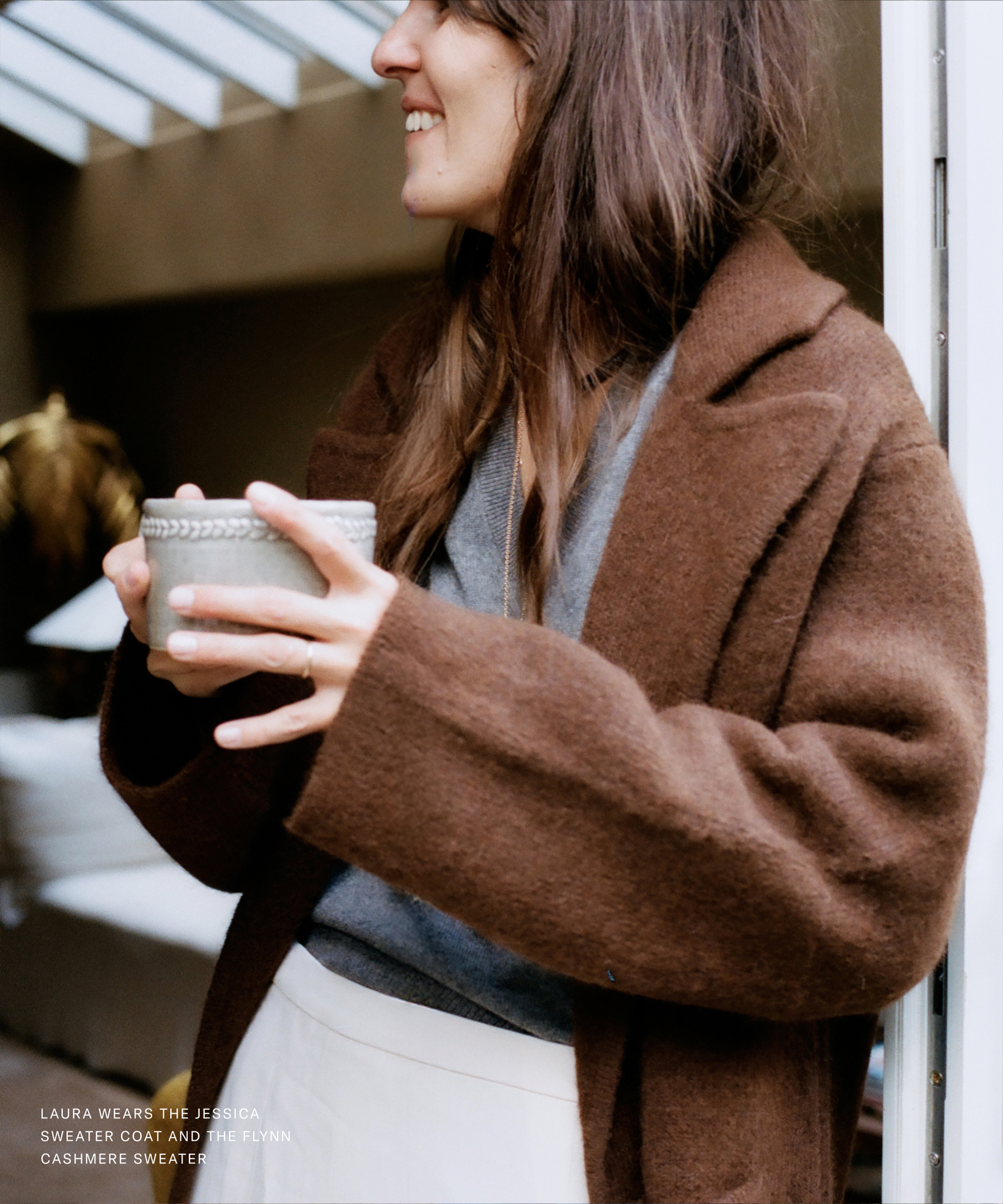 A woman with long brown hair, wearing a brown sweater coat and a grey sweater, smiles while holding a mug in both hands. The softly blurred background and text in the corner highlight Laura Vidrequin Roso's effortlessly chic outfit.