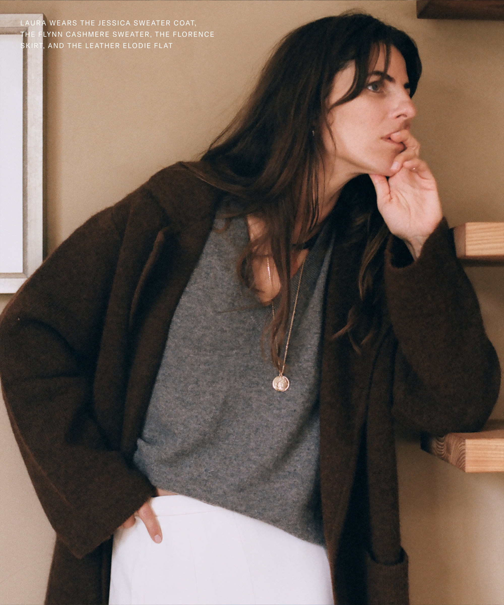 Laura Vidrequin Roso, with long brown hair, wearing a dark brown coat, gray sweater, and white skirt, leans against a wall with a thoughtful expression and hand on her chin. Shelves and text are visible in the background.