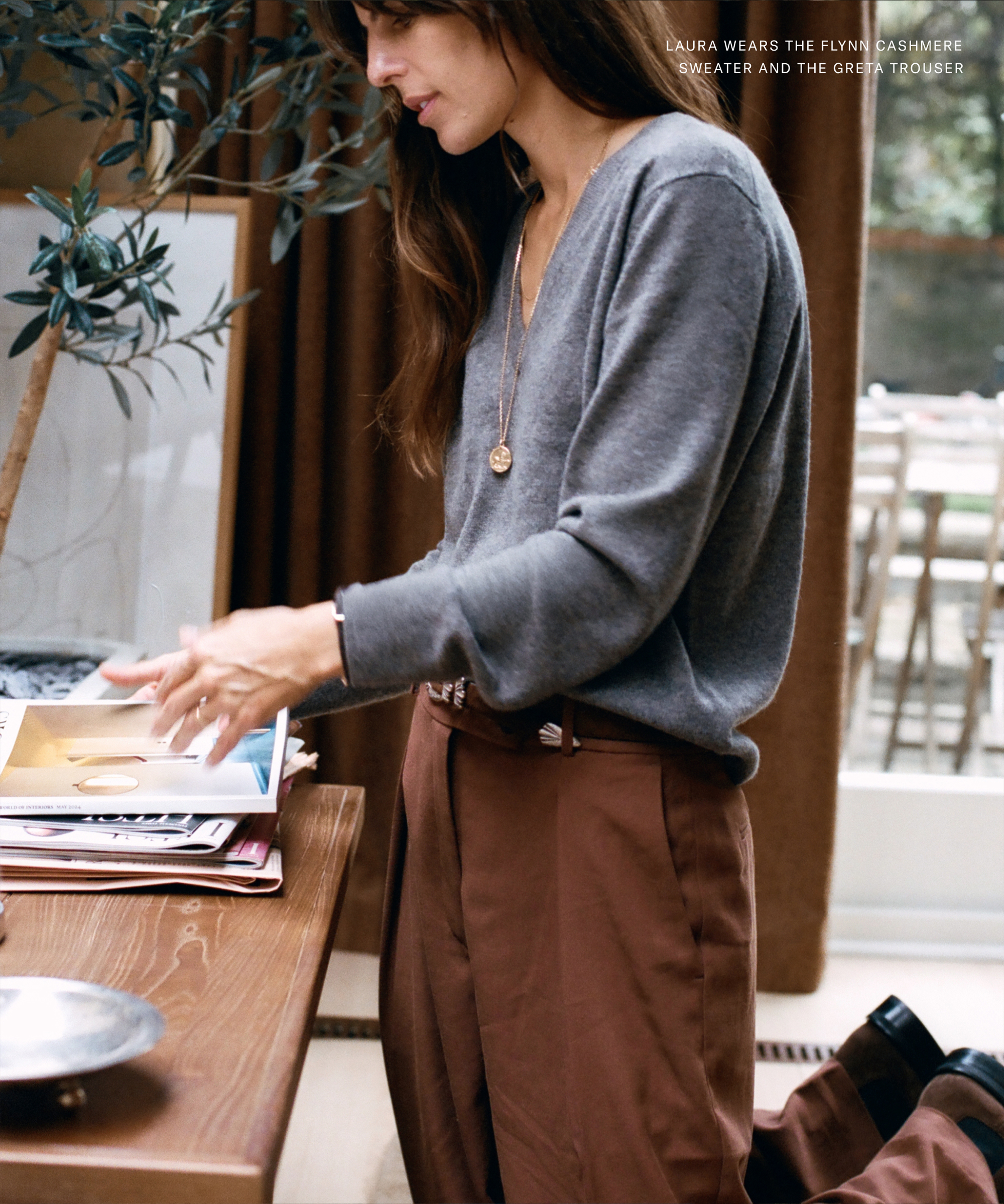 Laura Vidrequin Roso, with long brown hair, stands beside a table in a warmly lit room, wearing a grey cashmere sweater and brown trousers as she looks through papers near a potted plant and stacks of books.