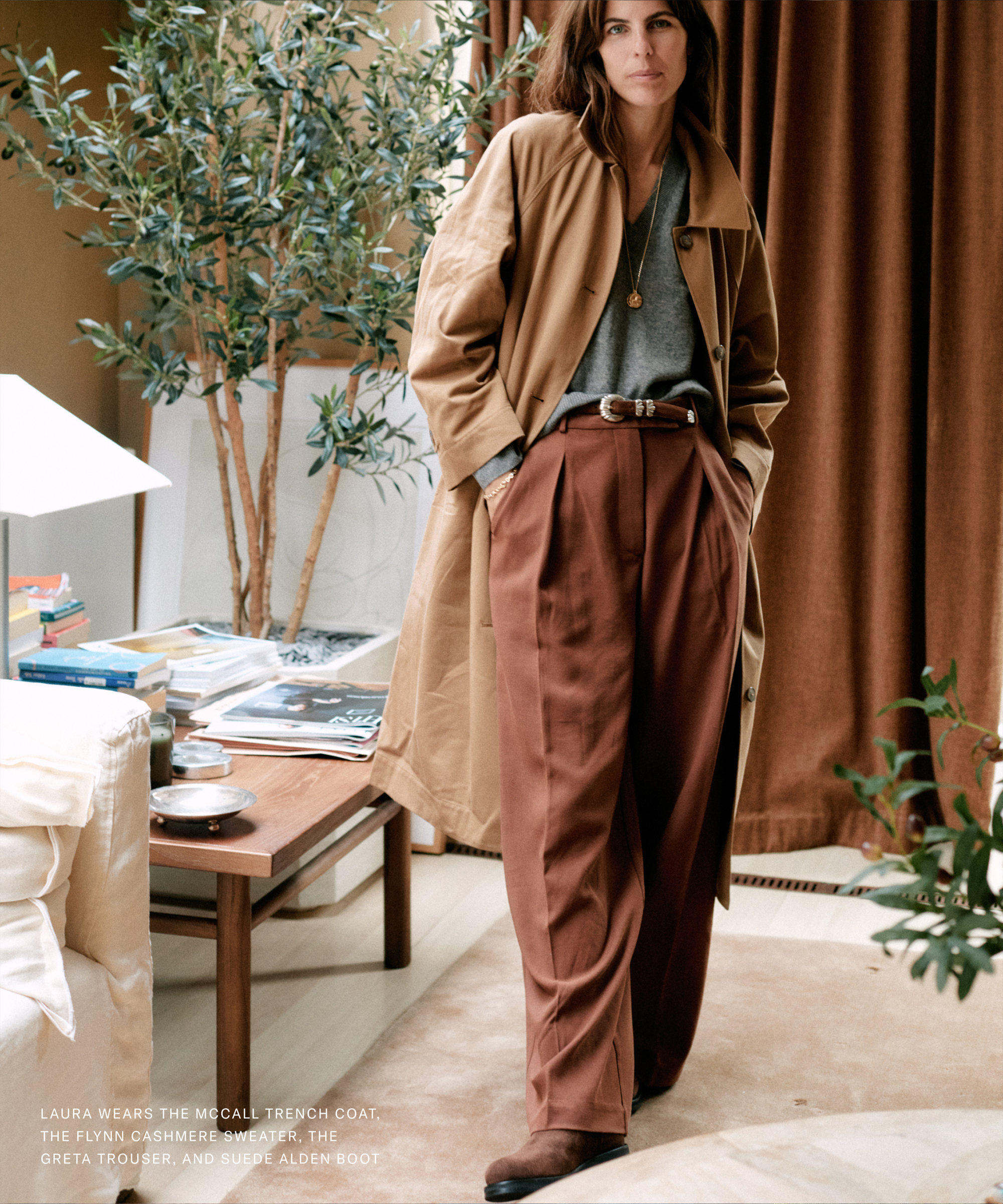 A woman stands indoors, wearing a tan trench coat over a gray top and high-waisted brown trousers. Inspired by Laura Vidrequin Roso's effortless style, she leans against a table with books, framed by a plant and brown curtains in the background.