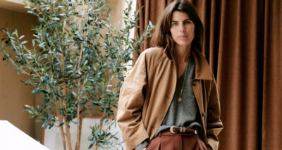 Laura Vidrequin Roso stands indoors near a leafy plant and brown curtains, wearing a tan jacket over a gray top and brown trousers, accessorized with a belt and a necklace.