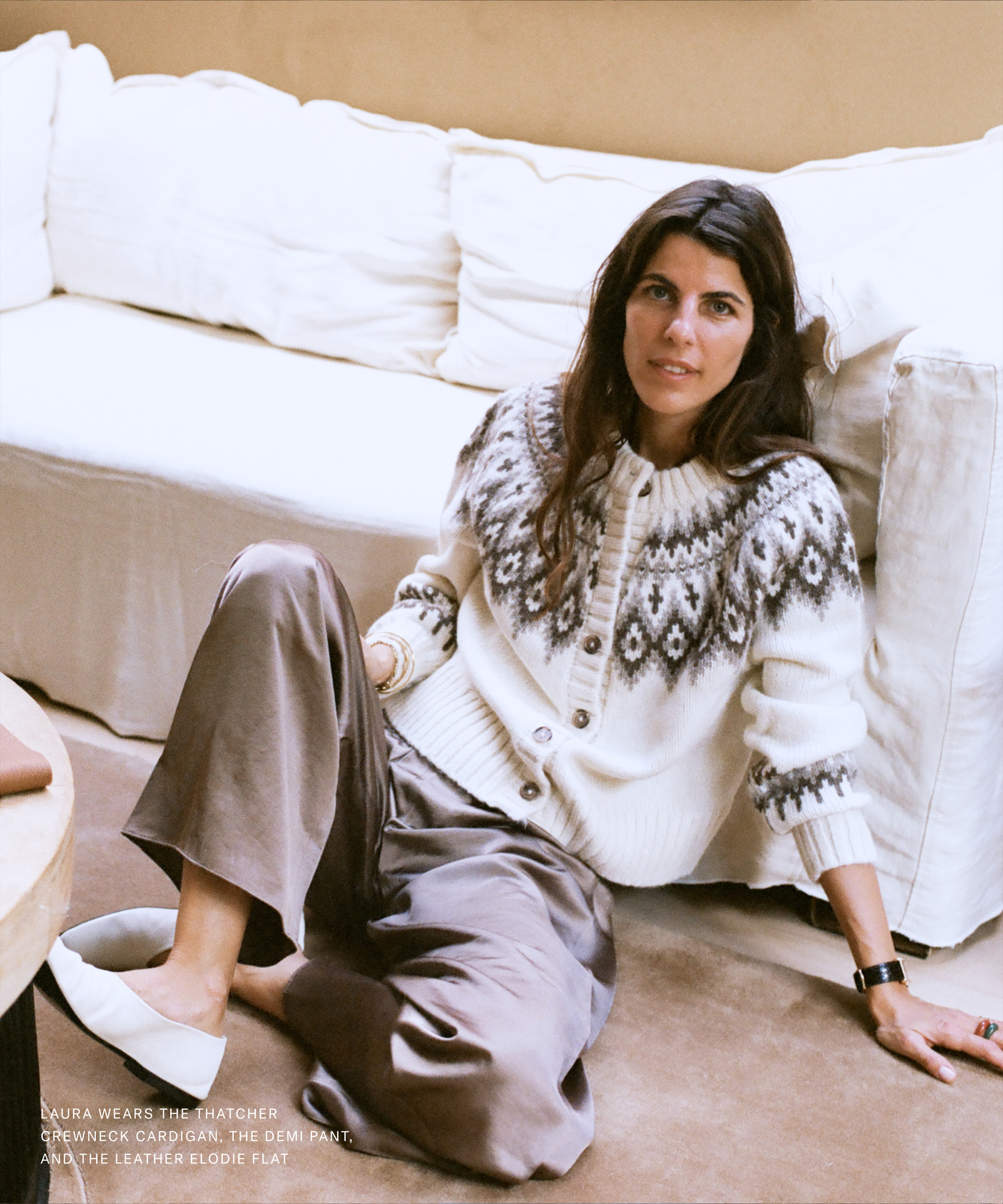 A woman with long dark hair sits on the floor beside a cream-colored sofa. Reminiscent of Laura Vidrequin Roso’s effortless style, she wears a patterned knit cardigan, wide-legged taupe pants, and white flats, exuding a relaxed confidence.