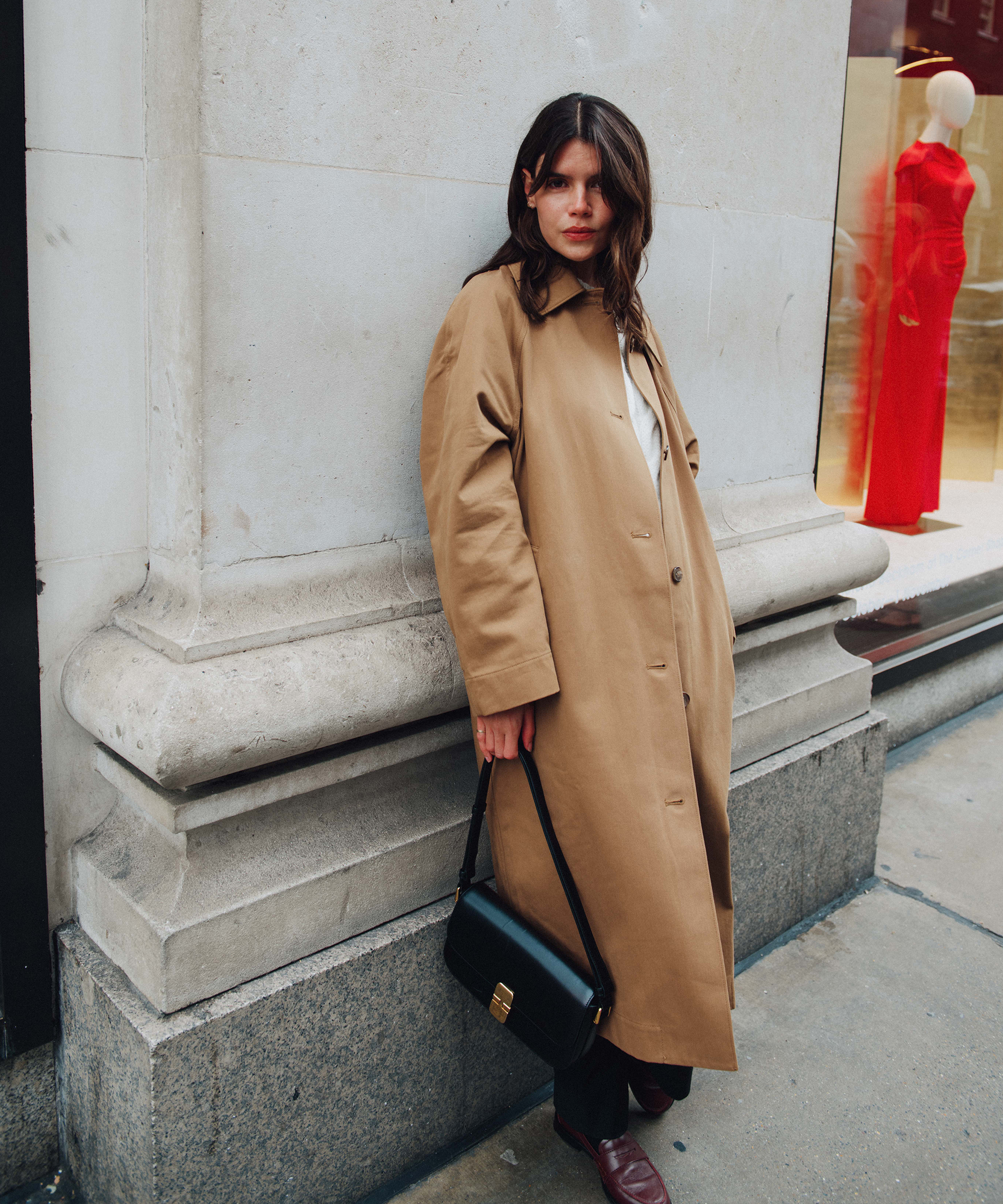 A woman with long brown hair wearing a tan overcoat, dark pants, and holding a black handbag stands against a stone building near Selfridges. Behind her, a shop window displays a mannequin in a bright red dress.