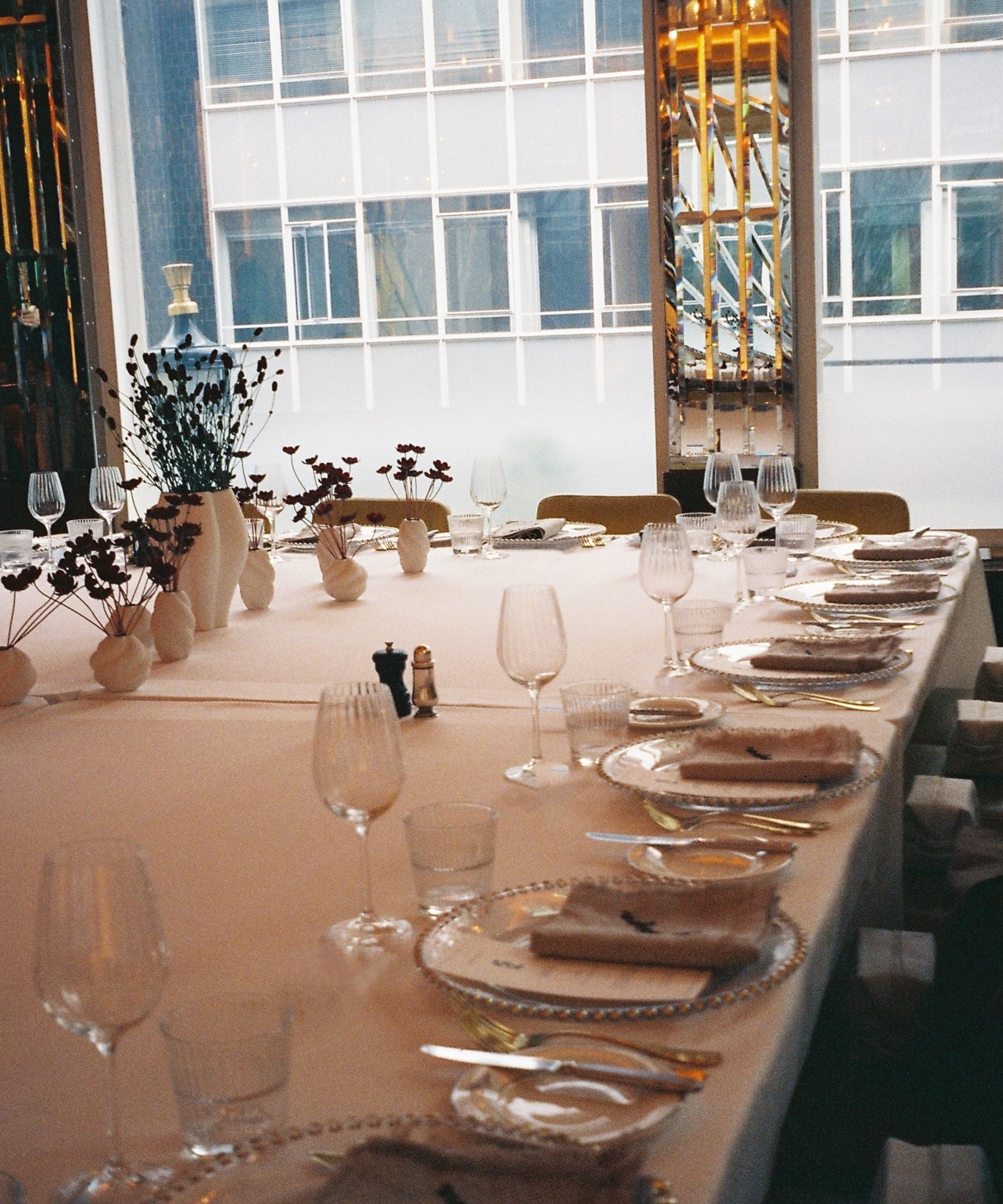 A long dining table set for a formal meal, with glassware, plates, folded napkins, and minimalist floral centerpieces in white vases. Large windows in the background reveal the modern Selfridges office building.