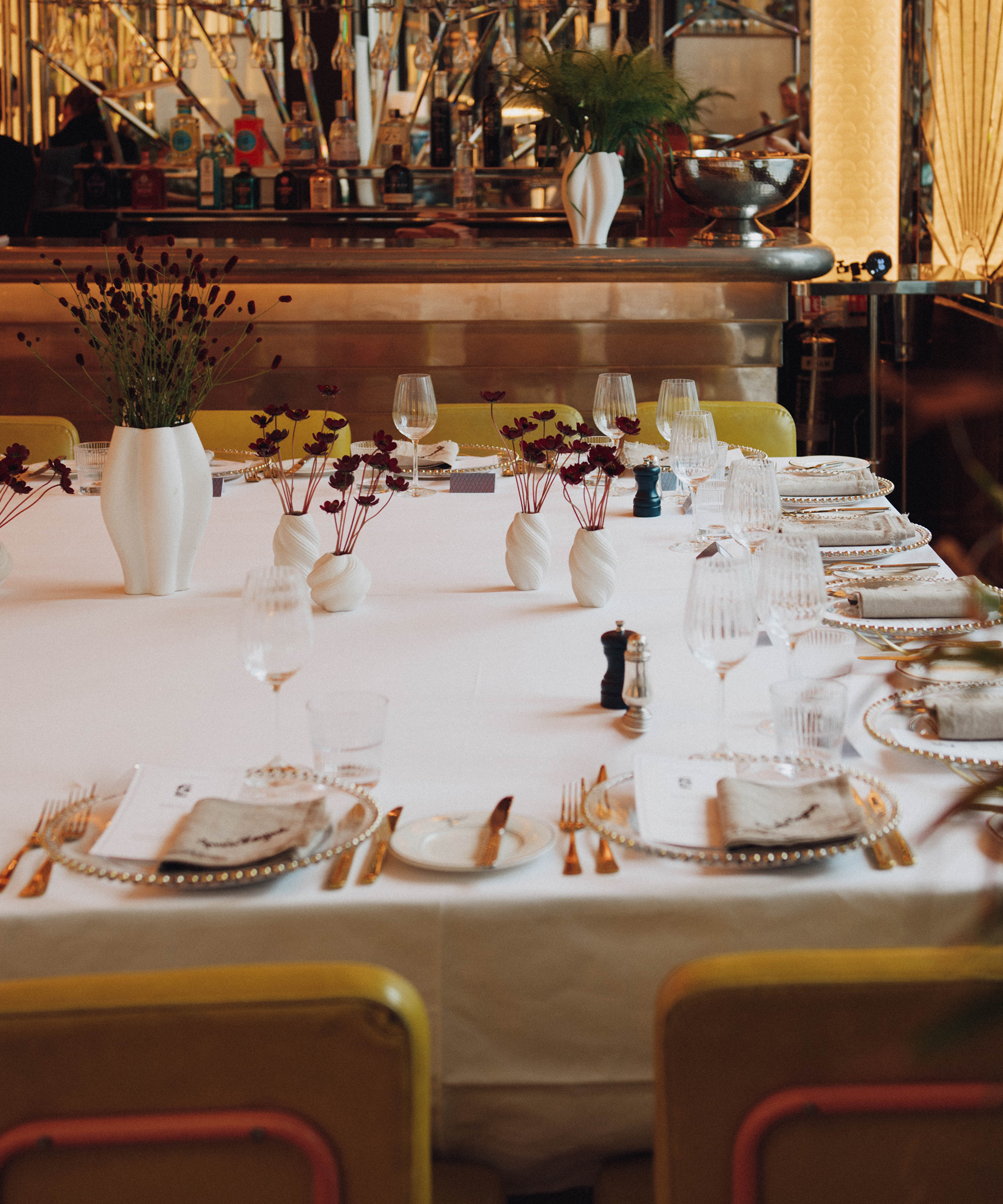 A large elegant dining table set with white linens, gold cutlery, clear glassware, and decorative white vases filled with dark red flowers. The background shows a Selfridges bar area and warm ambient lighting.