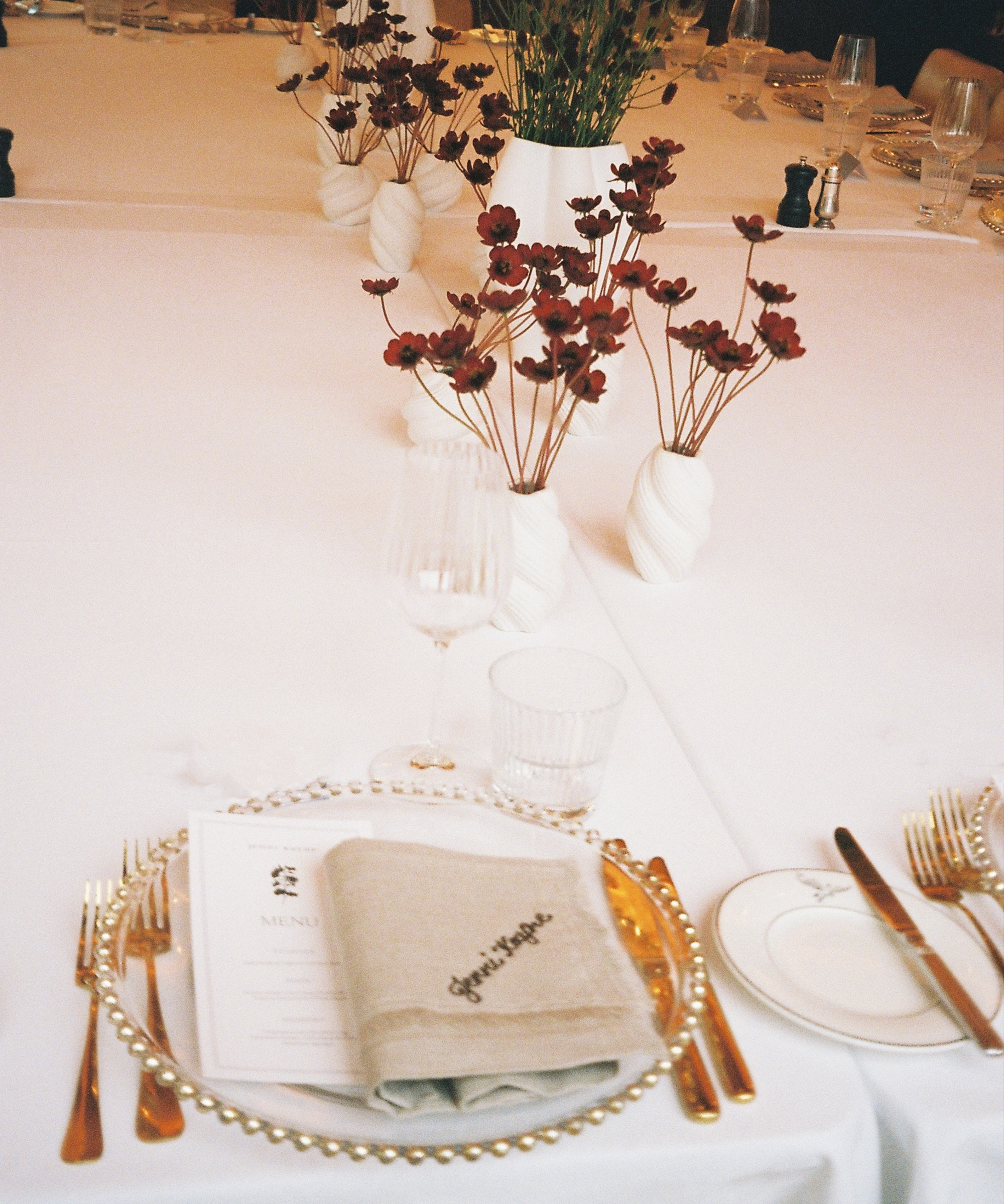A formal table setting with gold cutlery, a glass charger plate, a menu, and a beige napkin embroidered with “Good Hope” evokes the refined elegance of Selfridges, complemented by simple floral centerpieces on a white tablecloth.
