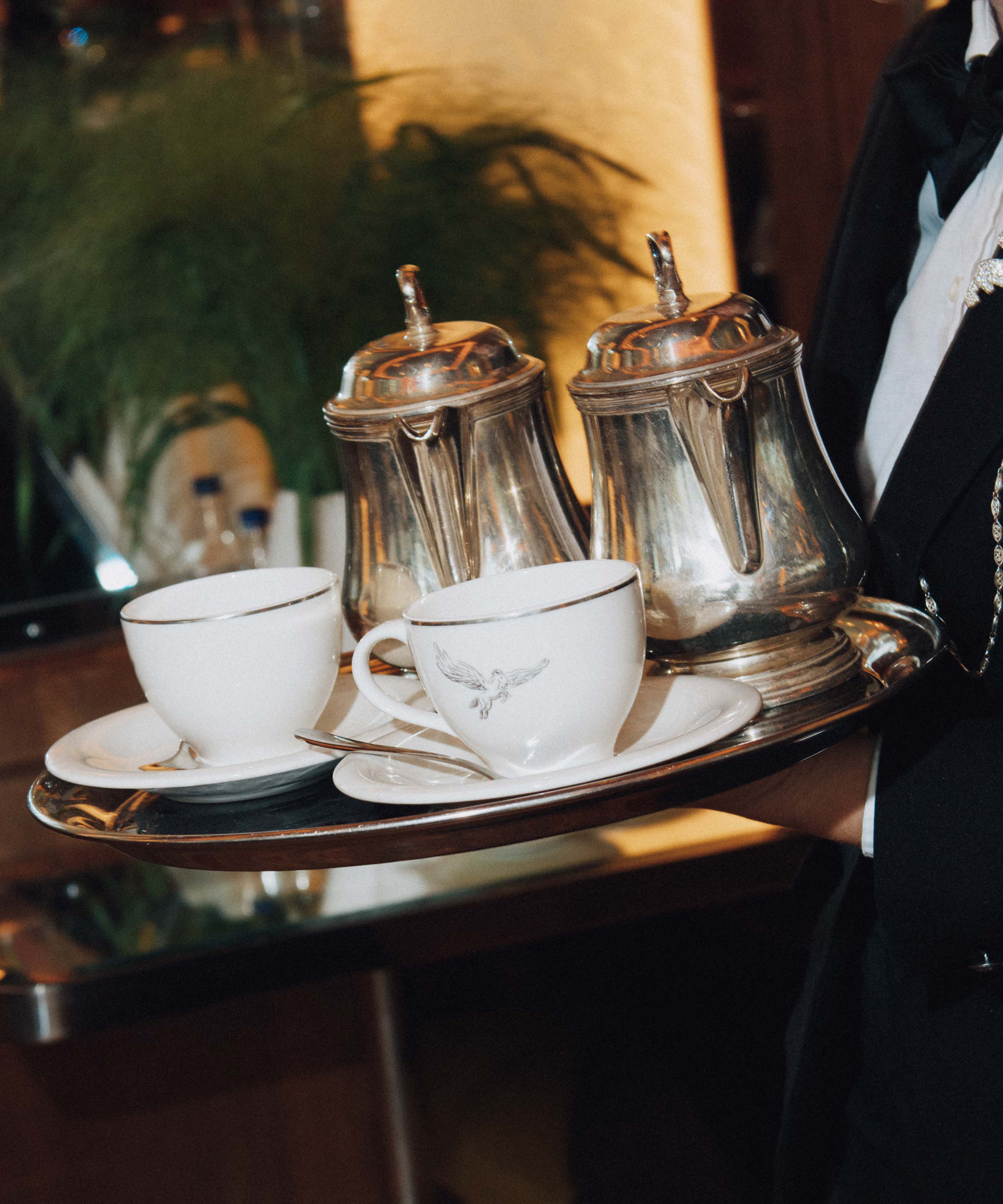 A server at Selfridges holds a silver tray with two white teacups, saucers, and ornate silver teapots. The background is softly blurred, filled with greenery and warm lighting.