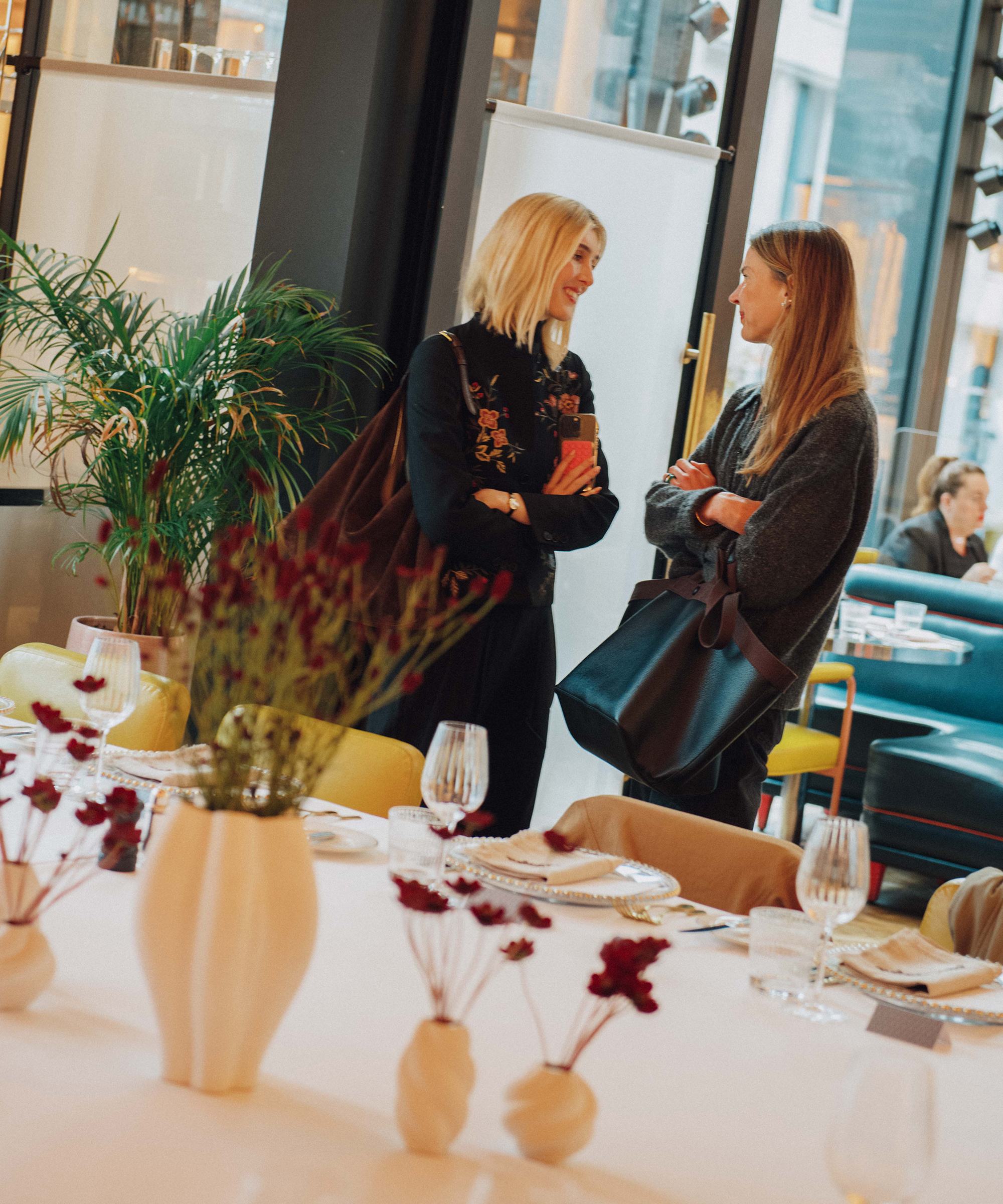 Two women stand and chat near a table set for a meal in a stylish restaurant at Selfridges. The table features white vases with red flowers, glassware, and plates. Large windows and modern decor are visible in the background.