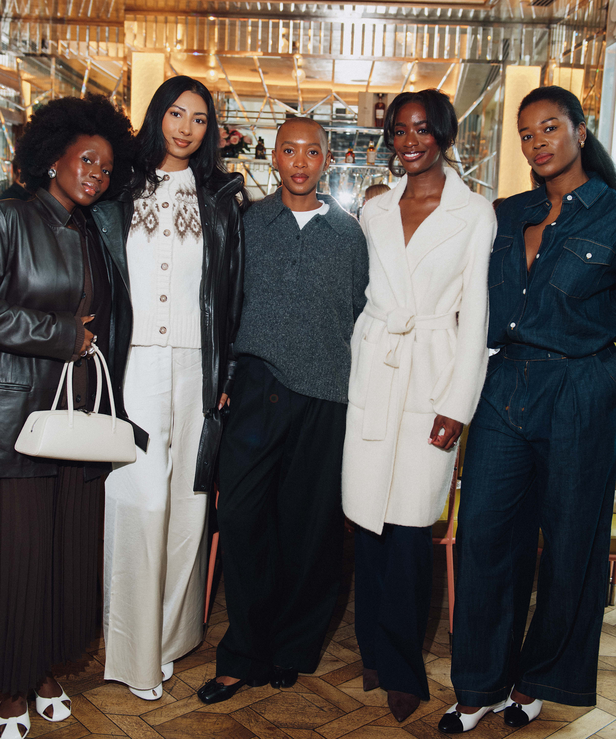 Five stylish women stand together indoors at Selfridges, dressed in fashionable outfits including leather, knitwear, a white coat, and a denim jumpsuit. They smile and pose in a warmly-lit, elegant setting with mirrored walls.