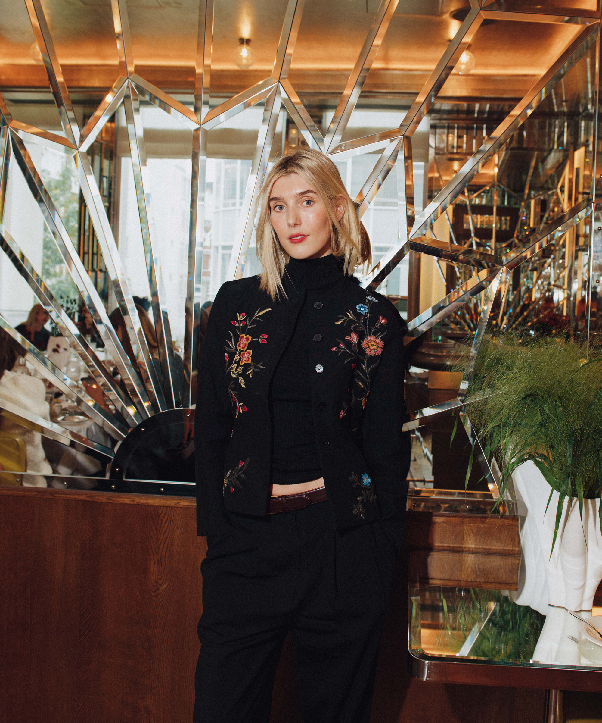 A woman with blonde hair wearing a black embroidered jacket and black pants stands in front of a mirrored, geometric wall at Selfridges. She poses with one hand in her pocket, next to a green plant in a white vase.
